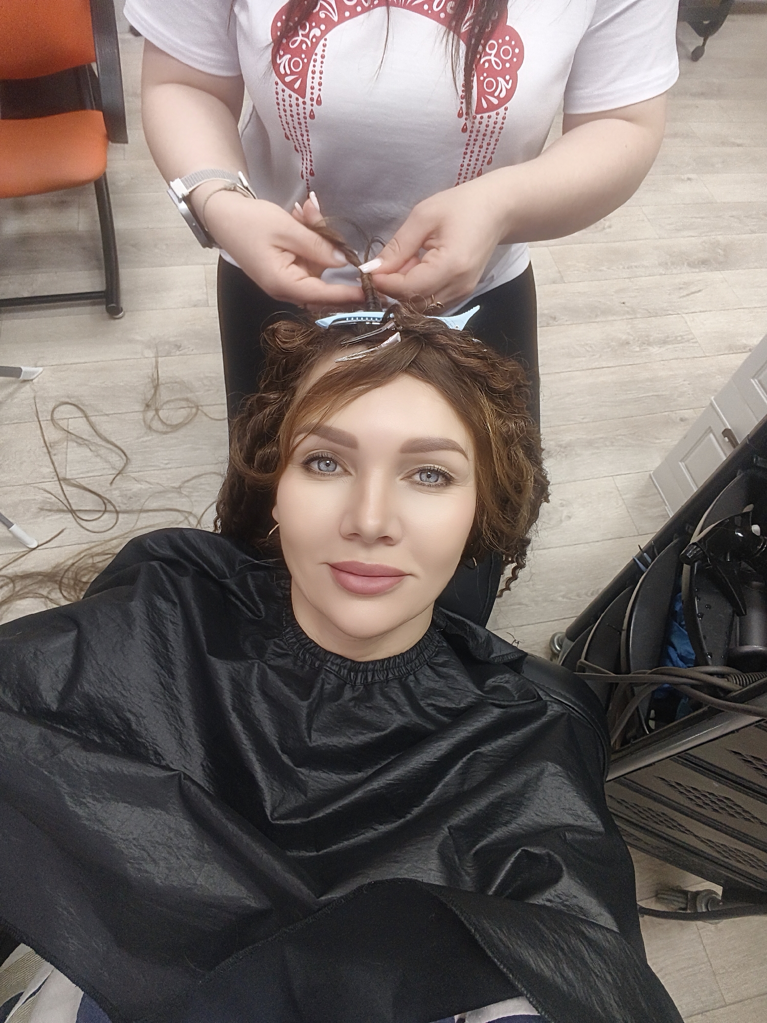 The image captures an indoor scene within what appears to be a hair salon, focusing on a client receiving hair styling services. In the foreground, a woman is seated, looking directly at the camera with blue eyes and a neutral to slightly pleasant expression. She has light brown, curly hair, with several sections on top of her head held by blue and white clips, suggesting a curling or perming process. She is wearing a black, synthetic protective cape around her shoulders.
Behind her, a hairdresser is actively working on her hair. Only the hairdresser's hands and upper torso are visible. They are wearing a white short-sleeved t-shirt with a red patterned design and a watch on their left wrist. The hairdresser's hands are seen holding and twisting a section of the client's hair, indicating careful styling.
The setting features a light-colored wooden floor with several strands of cut hair scattered on it. To the right, a black salon trolley or cart is visible, laden with tools such as a spray bottle and tangled electrical cords. A glimpse of an orange chair can be seen in the background on the left side of the frame. There is no discernible text visible in the image.