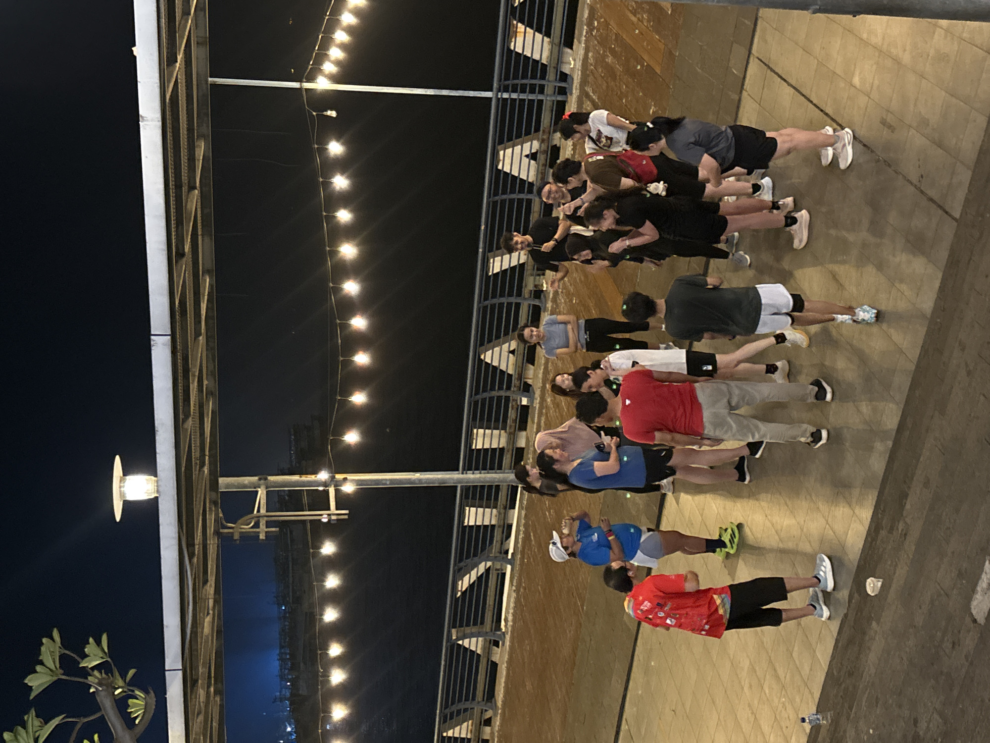 A group of approximately 15 adults, dressed in athletic attire, are gathered in a circle on a tiled outdoor area at night. They appear to be participating in an organized activity, possibly a warm-up or briefing, with some individuals facing inwards towards a person holding a microphone. String lights are visible overhead, illuminating the scene. The background features a dark body of water and a railing along a walkway. The location is identified as North Jakarta, Indonesia.
