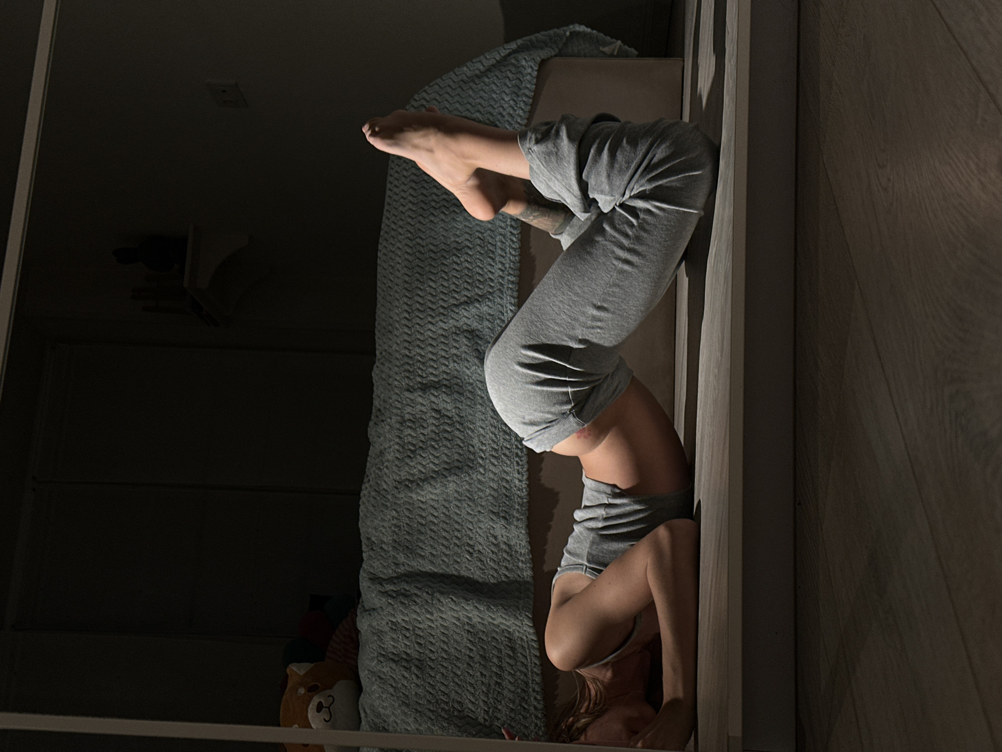 A person is lying on the floor in a dimly lit room. They are wearing grey sweatpants and a grey tank top. Their body is arched, with their feet pointed towards the ceiling. A bed with a textured blue blanket is visible in the background. There is also a stuffed animal on the bed and a shelf on the wall with some objects. The floor is made of wood. The image appears to be a photograph taken indoors. There are no explicit visual cues in the image that indicate the location is Moscow, Russia.