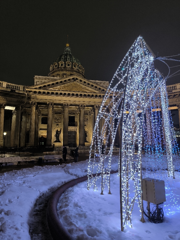 The image presents a nighttime winter scene in Saint Petersburg, Russia, centered on the Kazan Cathedral. In the foreground, a large, abstract decorative structure formed by a metal framework adorned with numerous small white and blue LED lights stands on snow-covered ground. This structure has a conical or arch-like shape. At its base, a beige electrical box with exposed dark wiring is visible. A low, curved, reddish-brown barrier or border delineates a pathway or boundary in the snow.
In the midground and background, the Kazan Cathedral dominates the left side of the frame. Its classical facade, constructed from light-colored stone, is illuminated by warm artificial lights. The building features a prominent portico with numerous tall columns supporting a triangular pediment, which bears a golden decorative element. Above the main structure rises a large, dark green dome topped with a slender golden spire, set against a dark, overcast sky. Snow lightly covers ledges and decorative elements of the cathedral. Beneath the main portico, two large, dark statues are visible in niches between columns.
Several indistinct human figures are present near the cathedral's entrance, appearing as dark silhouettes against the illuminated facade. They are standing on pathways or cleared areas amidst the snow. White park benches are partially visible, covered in snow, to the left of the illuminated structure. The ground throughout the scene is covered in a layer of white snow. The overall environment is dark and cold, with artificial lighting providing the primary illumination.