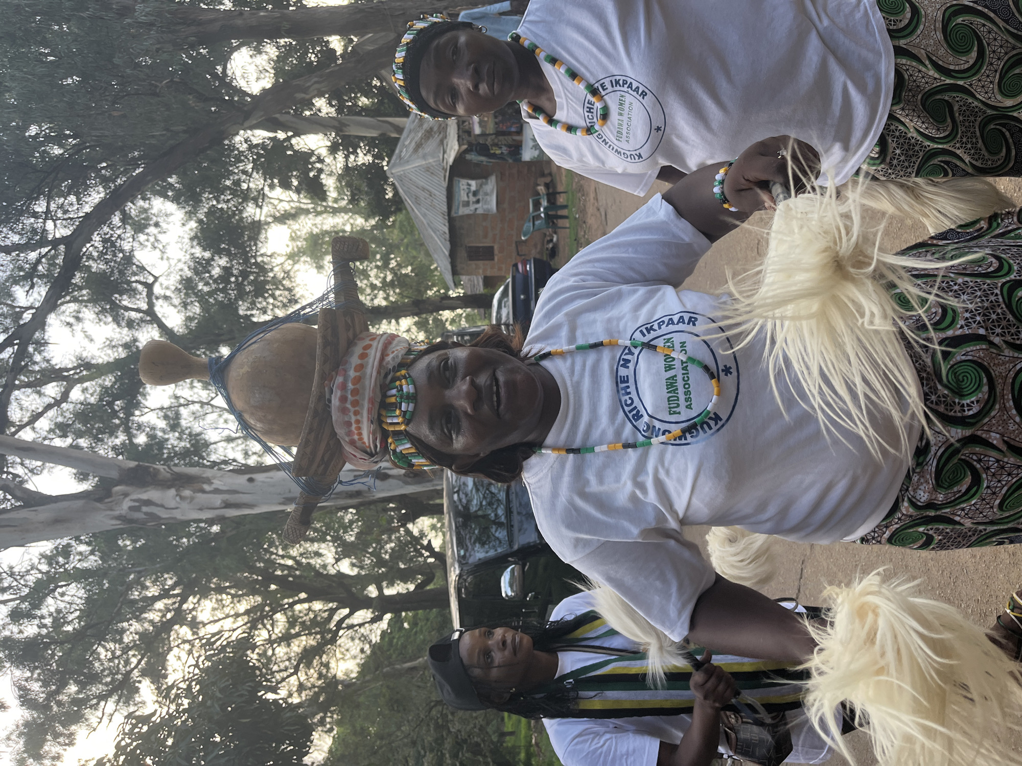 In an outdoor setting that appears to be in Jos, Nigeria, three women are the central focus of the image. The woman in the foreground, who is facing the camera, wears a white t-shirt with green and black lettering that reads "KUCHAN RICHE FUDAWA WOMEN ASSOCIATION". She has a beaded necklace around her neck, featuring green, orange, and white beads. Her hair is styled in cornrows, and she has a beaded headband with similar colors. She is holding a decorative object with long, white, fibrous strands.
To her left, another woman, also wearing a white t-shirt with the same "FUDAWA WOMEN ASSOCIATION" logo, is partially visible. She has her hair braided and is wearing a black baseball cap. She is also holding a similar decorative object.
To the right of the foreground woman, a third woman is visible from the chest up. She is also wearing a white t-shirt with the "FUDAWA WOMEN ASSOCIATION" logo and a beaded necklace. She has a beaded headband and is looking directly at the camera.
In the background, trees line the scene. A black SUV is parked behind the women, and a small, simple building with a corrugated metal roof is visible to the right. The overall lighting suggests it is late afternoon or early morning. The women appear to be participating in a cultural event or procession.