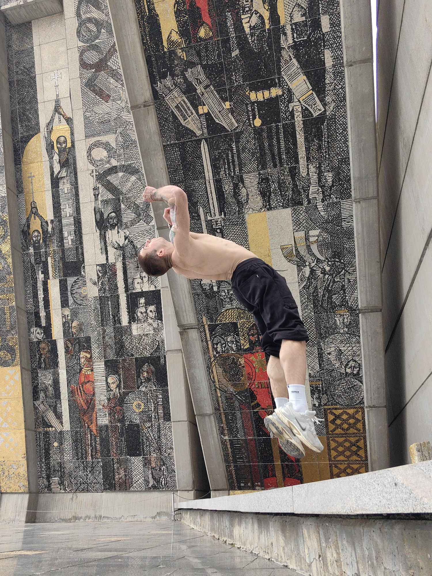 A man is captured mid-backflip in Shumen, Bulgaria. He is shirtless and wearing black shorts, white socks with "PAPER" printed on them, and worn white Nike sneakers. He is positioned on a gray concrete ledge. The background features a large, ornate mosaic mural in shades of gray, gold, red, and black depicting figures and religious scenes, with visible joints marking individual tiles. The mural extends vertically with sections partitioned by concrete pillars. The location appears to be a public space, likely near the "Founders of the Bulgarian State" monument, noted for its prominent mosaics.