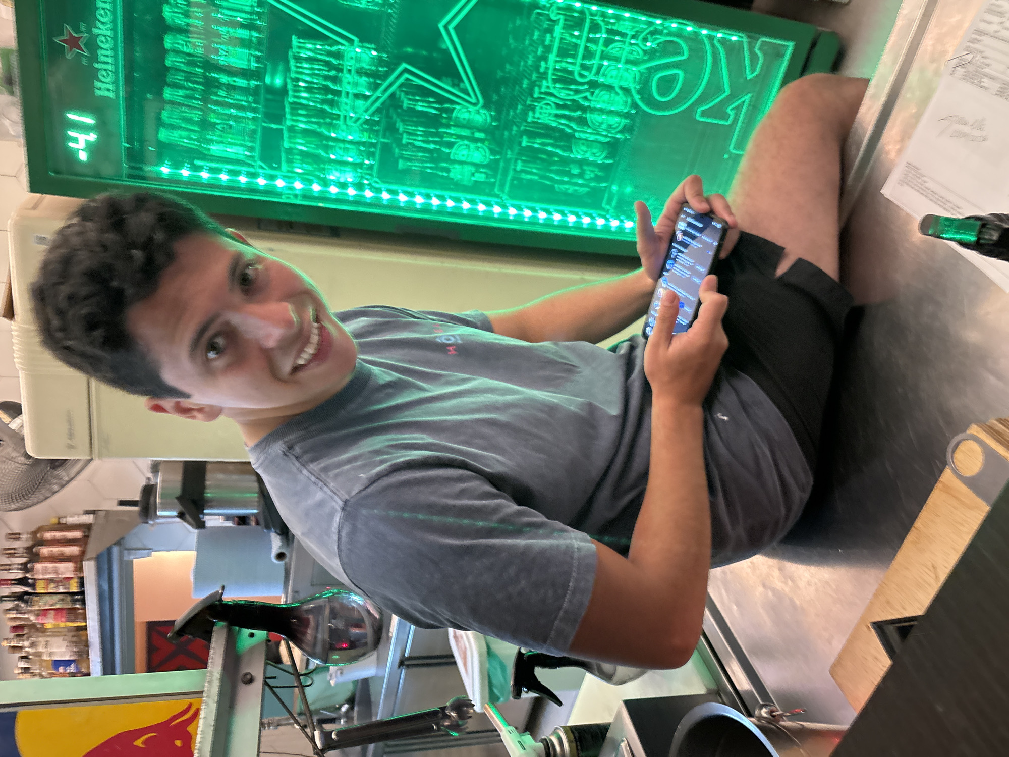 The image shows a young man with dark hair sitting at a bar. He is wearing a gray t-shirt and black shorts. He is holding a smartphone in his hands and looking at the camera with a smile. Behind him is a green neon sign with the word "Heineken" on it. The bar appears to be well-stocked with various bottles of alcohol. There is also a tap system and a cutting board on the counter. Based on the style of the bar and the general atmosphere, it's possible that this image was taken in the city of São Paulo, Brazil.