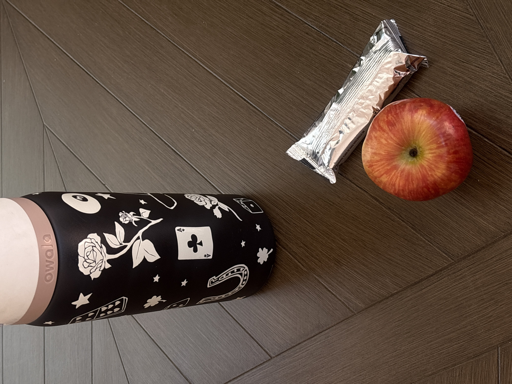 The image captures an overhead view of a water bottle, an apple, and a foil-wrapped snack bar laid out on a dark brown wooden floor. The flooring features a distinctive herringbone or chevron pattern.
The water bottle, identified as an Owala brand, is black with numerous white outline illustrations. These designs depict various symbols, including roses, an eight-ball, stars, dominoes, a horseshoe, a four-leaf clover, an ace of clubs, and dice, giving it a thematic look. A light pinkish-beige band near the top of the bottle clearly displays "owala" in white text.
Adjacent to the bottle, a fresh red and yellow striped apple rests, its stem end facing upwards. Beside the apple, a rectangular snack bar is visible, enclosed in crinkled silver foil packaging. Partial text, such as "SADO NO SOST" and "PARA SU VENTA," can be discerned on the wrapper, likely indicating product information or instructions.
The arrangement of these items suggests a prepared snack or part of a light meal. The scene is static, implying readiness for consumption rather than active preparation or eating. The setting is indoors, with ambient lighting that does not indicate a specific time of day. The context for this scene is Puebla, Mexico.