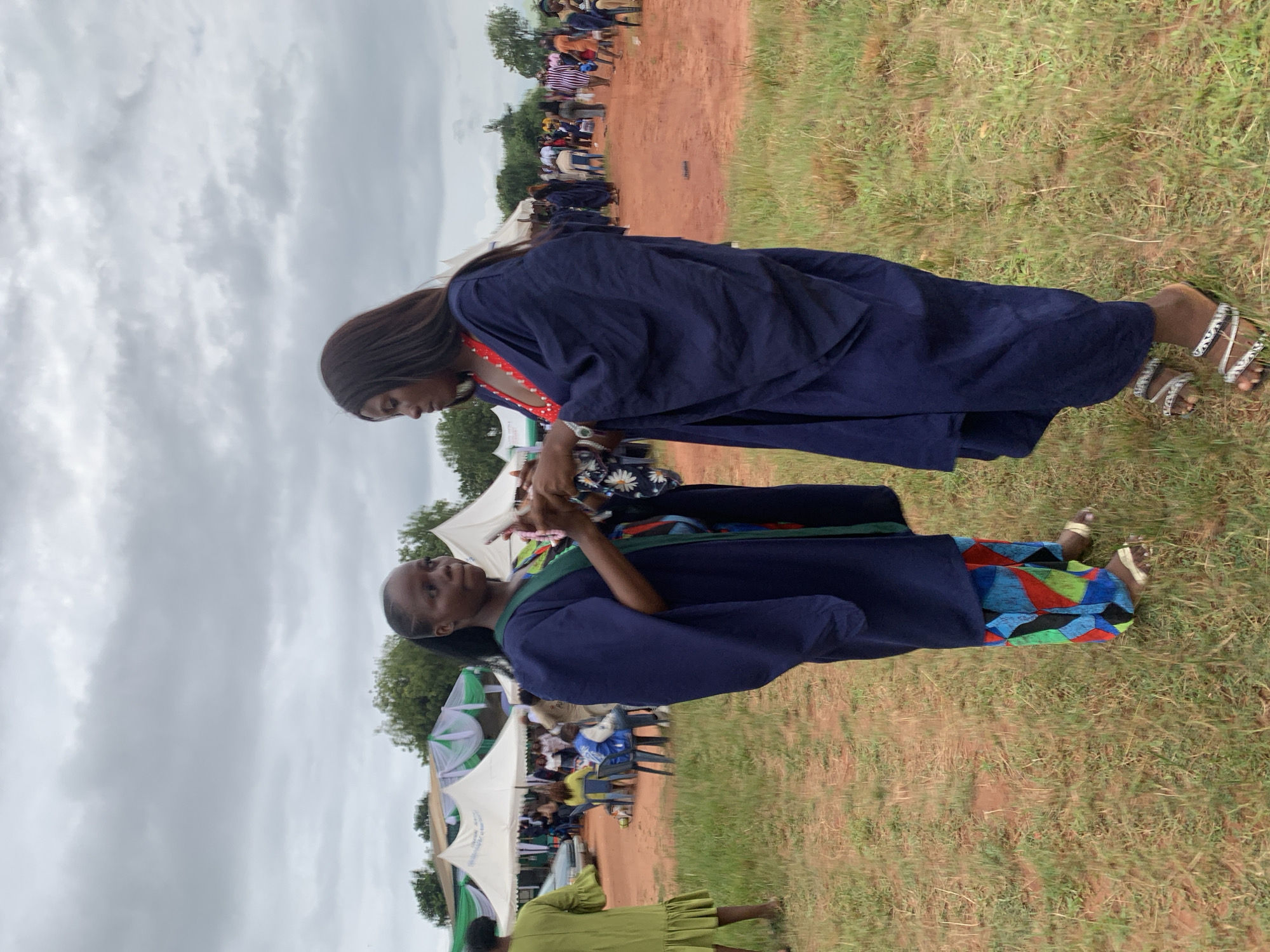 Two women are standing face-to-face in an outdoor setting. Both are wearing dark blue graduation robes. One woman has long black hair and a red and white patterned collar. The other woman has short hair and a green collar; her robe is decorated with red, green, and blue patterns. In the background are tents and people, suggesting a gathering or event. The location appears to be in Nsugbe, Nigeria, as the scene has the environmental characteristics of that area.