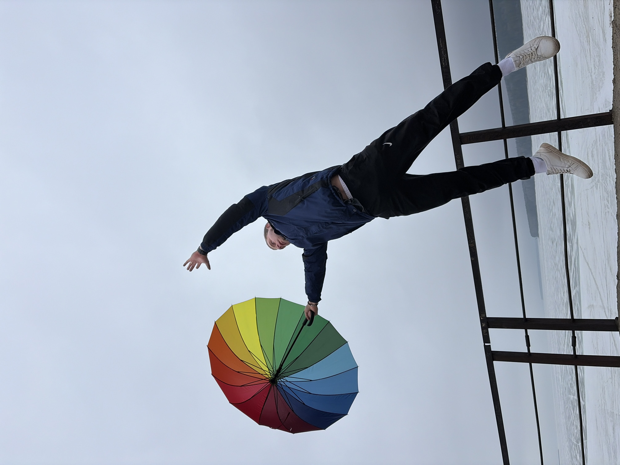 A man in a navy jacket and black track pants stands on a structure next to a water feature in what appears to be the city of Chaikovsky, Russia. He is holding a closed, rainbow-colored umbrella. His left arm is extended outwards. He is wearing white sneakers and socks. The background is a cloudy sky.