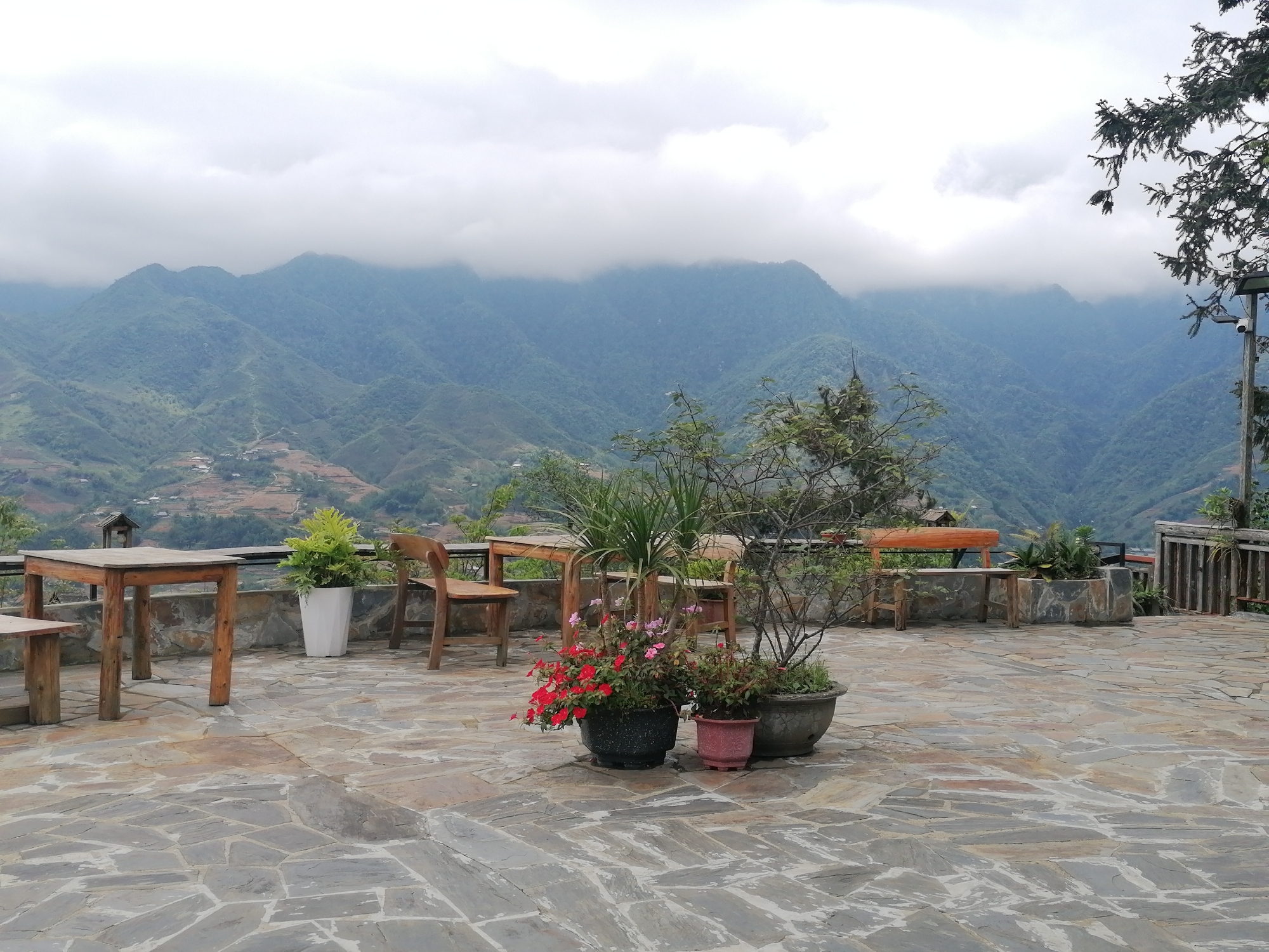 The image depicts an outdoor patio area in Sa Pa Ward, Vietnam, overlooking a mountainous landscape. The sky is overcast. The patio is paved with irregular stone slabs and furnished with wooden tables, chairs, and benches. Several potted plants are arranged around the patio. In the background, a series of green mountains are visible, some obscured by low-hanging clouds. A wooden fence borders a section of the patio.