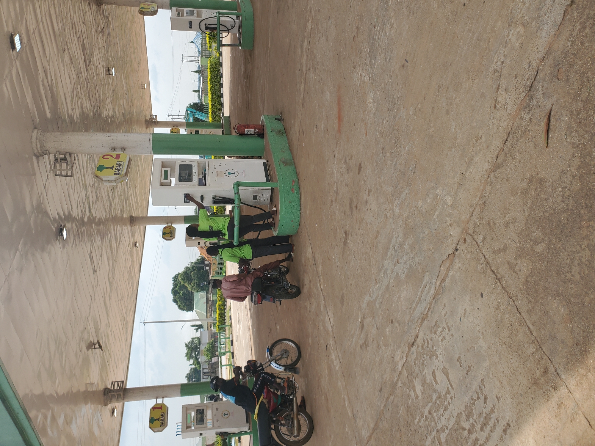 This image depicts an outdoor scene at a gas station, likely in the city of Winters, United States of America, though this specific location cannot be definitively confirmed from the image alone. The scene is dominated by a covered forecourt with concrete paving. Several white gas pumps are visible, some with green trim. One pump has a red fire extinguisher mounted to its side.
In the foreground, two motorcycles are parked. One is a red and black motorcycle ridden by a person wearing a black t-shirt and dark pants, who is partially obscured by the motorcycle. The other motorcycle is a dark color and is ridden by a person in a light brown traditional garment and a cap.
In the middle ground, two individuals in bright green shirts appear to be interacting with a gas pump. One is standing and facing the pump, while the other is seated on the motorcycle, also facing the pump. The green shirts suggest they may be employees of the gas station.
In the background, there are trees, buildings with various roofing styles, and utility poles, indicating an urban or semi-urban environment. The sky is partly cloudy. The overall impression is of a typical, functional gas station.