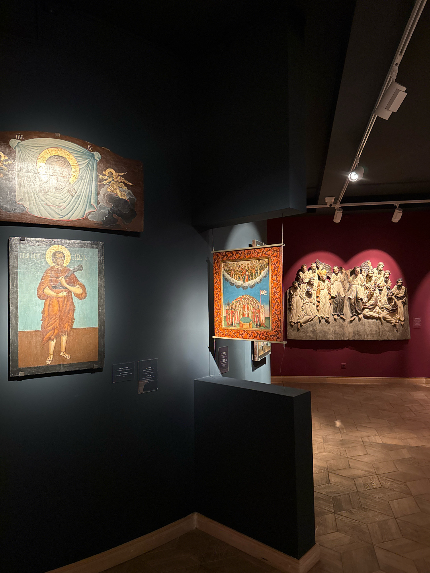 An indoor shot captures a museum exhibition featuring religious artwork. On a dark blue wall, two icons are displayed: one at the top depicts a figure with a halo and draped cloth, with an angel to the right. Below it, a second icon shows a figure in an orange robe holding a staff or cross, standing against a light blue background. To the right, a vibrant tapestry with a border and a scene of figures in the sky hangs. Adjacent to the tapestry, a large stone relief sculpture of many figures, likely angels or saints, is mounted on a burgundy wall. The floor is a wooden parquet with a geometric pattern. Lighting is provided by track lights on the ceiling.