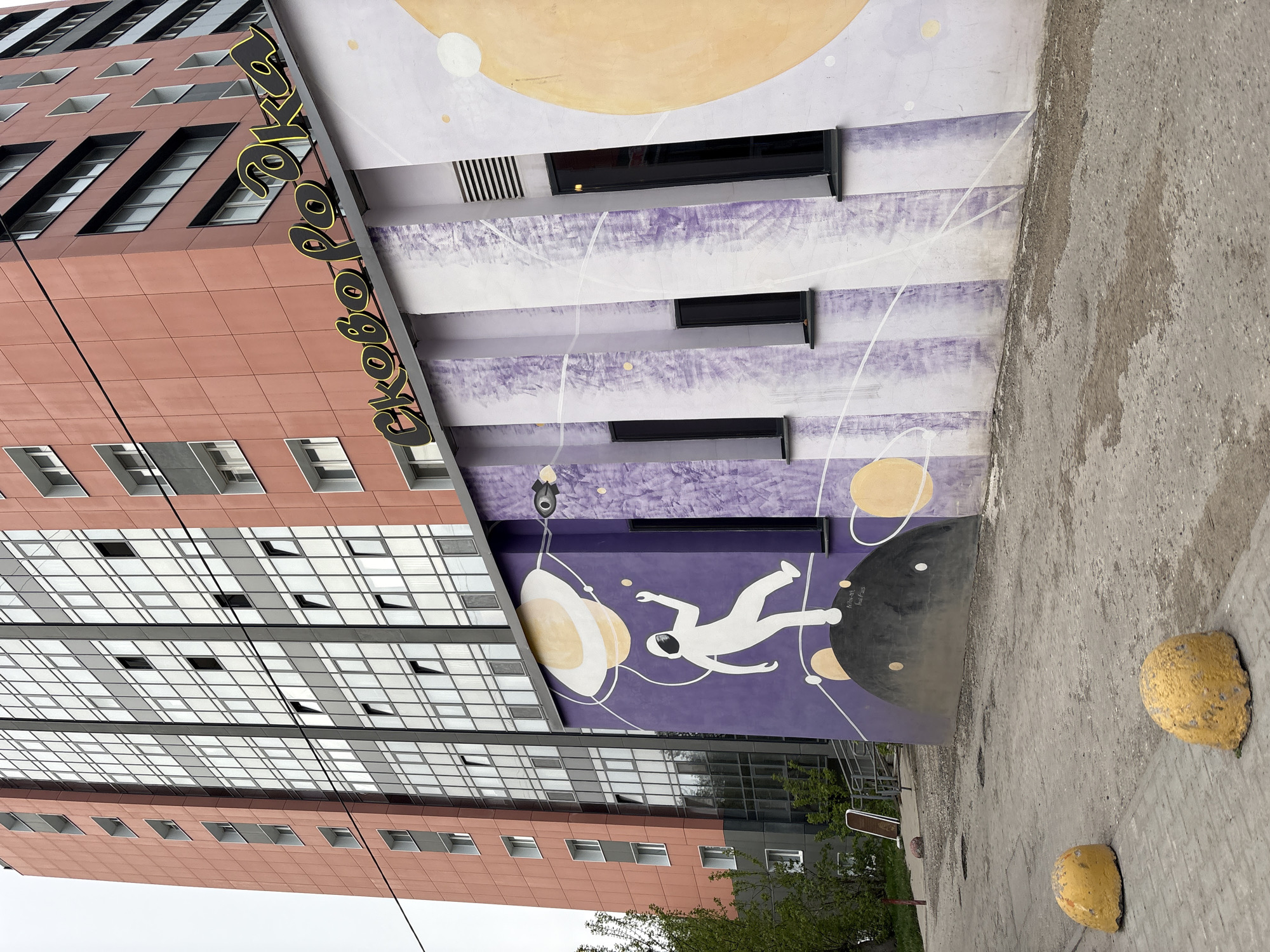 The image shows a tall building in Perm, Russia with a mural depicting an astronaut floating in space near planets. The mural is painted on a section of the building that is angled away from the main facade. The building has two distinct sections: one covered in glass windows and the other with a brown tile facade with smaller windows. The name of the building or business, written in Cyrillic, runs vertically along the edge where the two sections meet. In the foreground, there are two yellow, rounded bollards on a concrete surface.