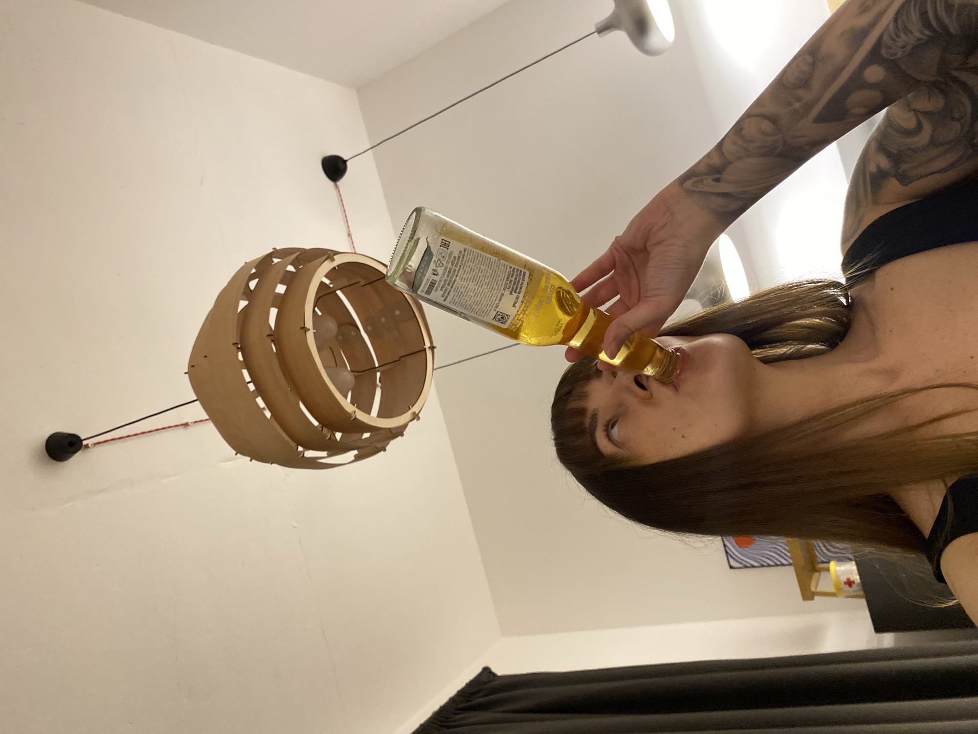 A woman with long brown hair is drinking from a beer bottle. She's wearing a black top. Her arm has a large tattoo. In the background, there's a wooden, circular lamp hanging from the ceiling. There's also a black curtain and a shelf with items on it. The image's location is Izhevsk, Russia.