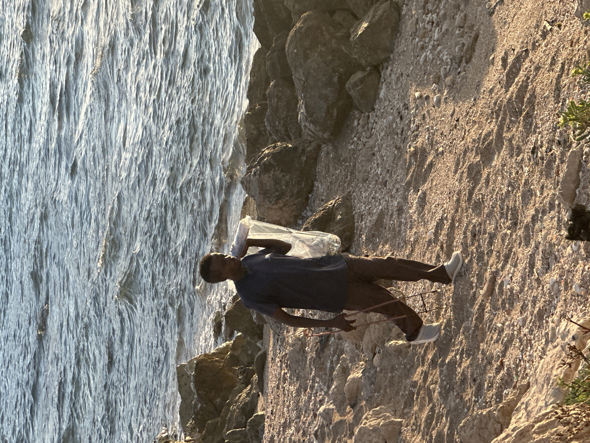 A young man stands on a rocky shoreline, holding a clear plastic bag over his shoulder and a pink, coiled object in his right hand. He is wearing a dark blue t-shirt and brown pants, with light-colored shoes. The scene appears to be a beach or coastal area, with the sea on the left and a rocky, sandy slope on the right. Large rocks are visible along the waterline and on the slope. The sun is casting long shadows, suggesting it is late afternoon or early evening. The water is choppy with light reflecting off its surface. The overall mood is casual and relaxed, with the man appearing to be engaged in some activity, possibly collecting or exploring. No visible text is present in the image. The location context provided indicates Haifa, Israel.