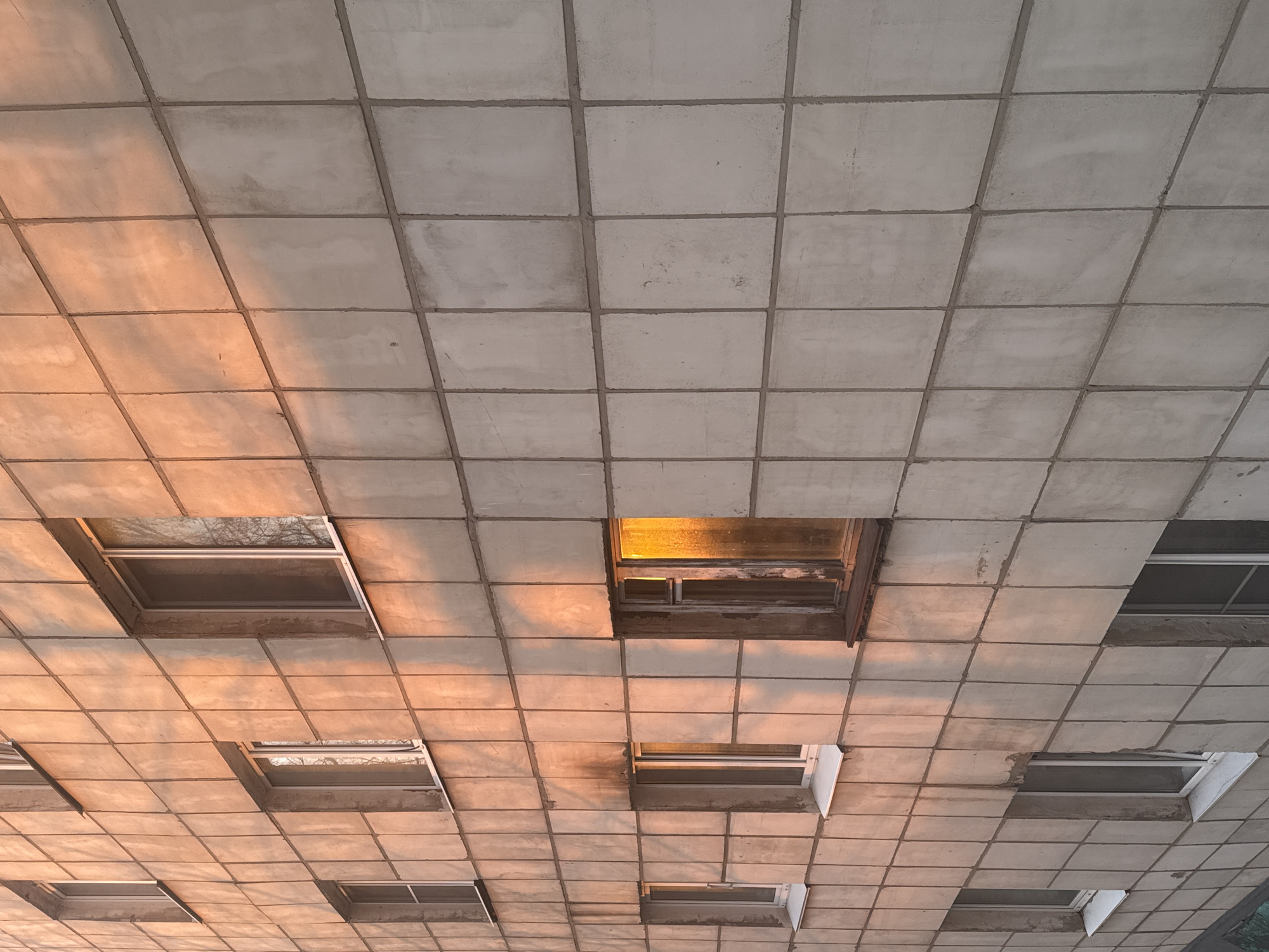 The image shows a low-angle view of a tiled ceiling with several square openings containing what appear to be windows. The ceiling is composed of light gray, square tiles with darker gray grout lines. Sunlight is casting an orange hue on the ceiling. The window frames within the openings are visible. The location is Unknown, Unknown. There are no people in the frame.