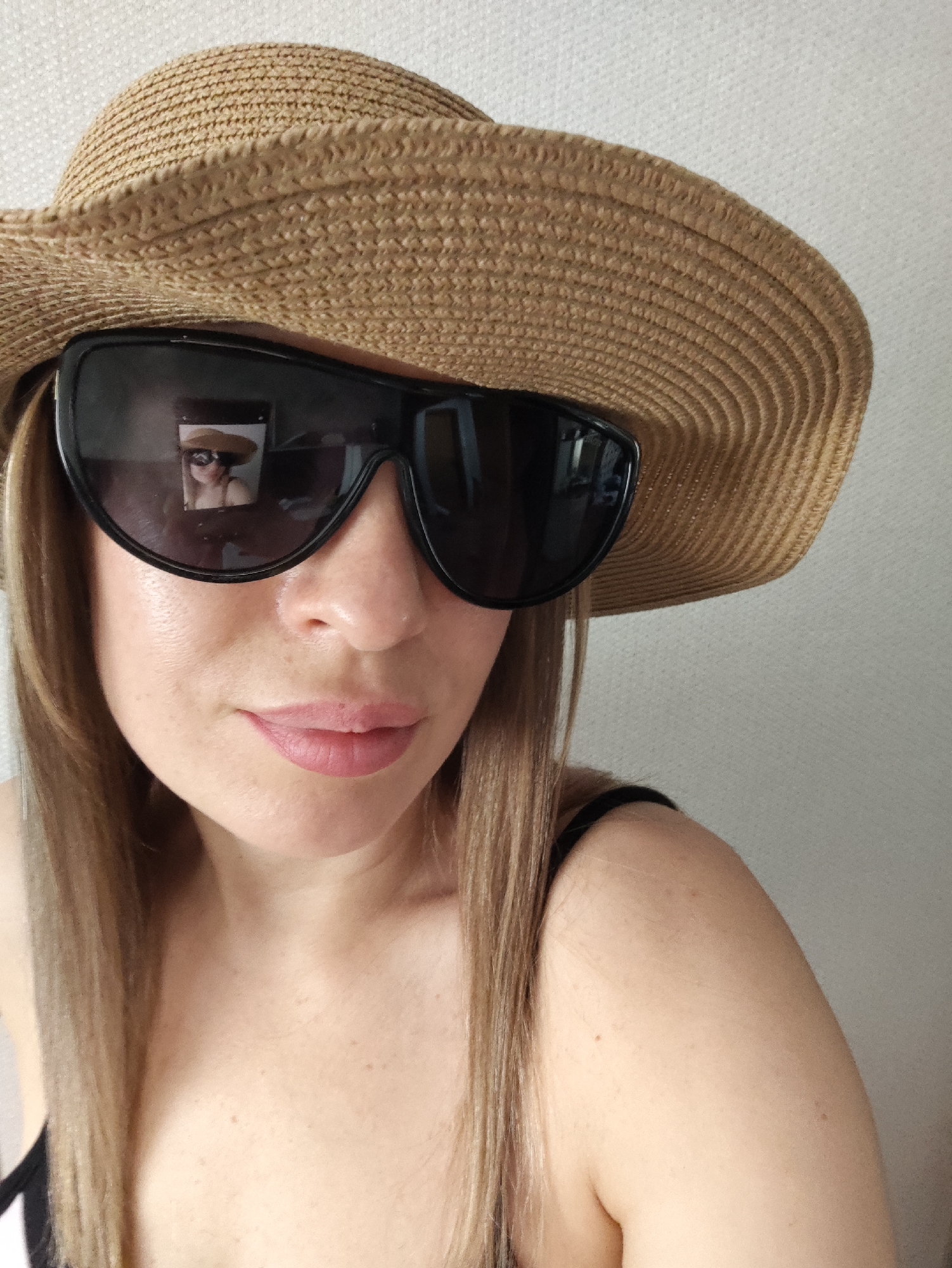 A close-up, selfie-style shot shows a fair-skinned woman with blonde hair, wearing a straw hat and large black sunglasses. Her lips are slightly parted, and she has a few faint freckles on her cheeks. The sunglasses reflect a small image of herself, also wearing the hat and sunglasses, with a blurred background. She is wearing a black tank top. The background appears to be a plain, textured wall. The image does not contain any discernible information that would allow for its location to be identified as Almaty, Kazakhstan.