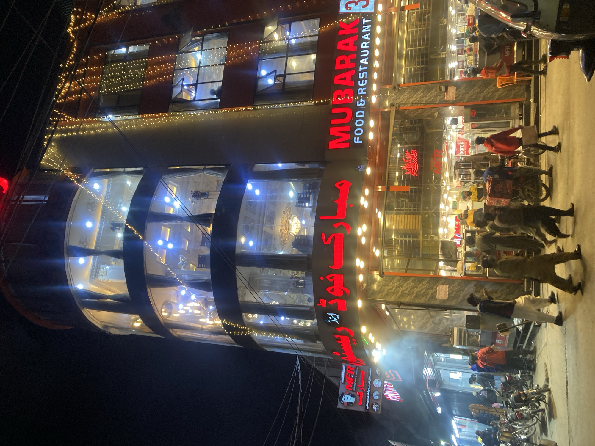 The image shows a multi-story restaurant named "Mubarak Food & Restaurant" at night, likely located in Quetta City Tehsil, Pakistan. The building is brightly lit with decorative lights outlining its architecture. Red signage displays the restaurant's name in both English and Arabic (possibly Urdu). People are gathered outside, some standing and others on motorcycles parked along the street. Delivery people are also visible. The environment is urban, with overhead power lines and surrounding buildings.