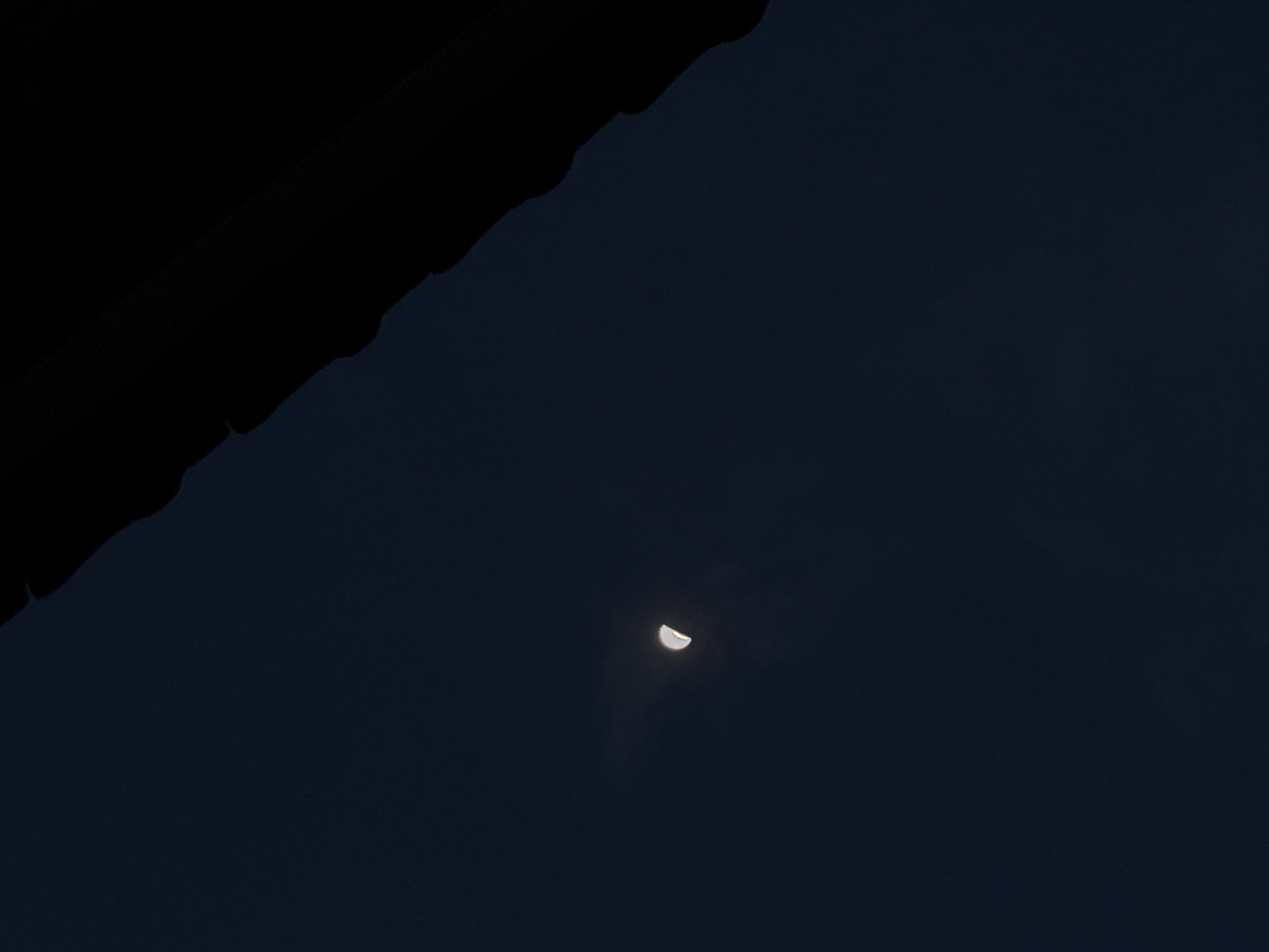 A low-angle, nighttime view of the sky, likely taken from Gadingrejo, Indonesia. The moon is in its crescent phase, positioned in the lower-center of the frame. In the upper-left corner, a dark, indeterminate object, possibly a roofline or tree branch, obscures part of the sky. The overall tone is dark and muted, with minimal detail visible beyond the moon and the foreground object.