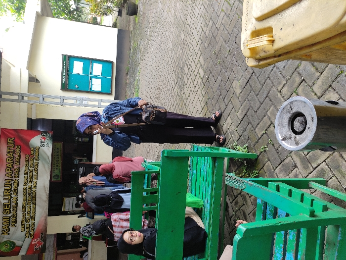 This image captures an outdoor waiting area, likely at a public office, in Kepanjen, Indonesia. The scene appears to be set during the day.

The primary subjects are several people, mostly women, some wearing hijabs. In the foreground, a woman in a black hijab and glasses is seated on a green wooden bench, looking towards the viewer. Slightly further back, a woman in a blue denim jacket, patterned blue hijab, and a cross-body bag stands and looks at her phone. Other individuals are visible in the background, mostly seated on similar green benches.

The setting includes a paved ground made of interlocking bricks. A light-colored building forms the background, with a green-framed notice board on its wall. To the right, part of a yellow plastic container (possibly a water tank or bin) and a metal ashtray or waste receptacle are visible.

The activity taking place appears to be people waiting. The woman standing is engrossed in her phone, while others are seated, suggesting a period of waiting or downtime.

A prominent red and white banner, resembling the colors of the Indonesian flag, is visible on the left side of the image. The text on this banner, as per the provided OCR, reads:
"KAMI SELURUH APARATUR
PENGADILAN AGAMA KAB, MALANG
BERTERED MEWUJIOKAN LOKA INMIGRITAD
MENUTU WILAYAH BEBAS DARU NORUPTI WERT DAN
PELAYANAN BIROKRASI BERSIH DAN MELAYANI (WBBM)"
Followed by "STP" and "DON PARTUKA VIKES". This text indicates the location is the Religious Court of Malang Regency and appears to be an integrity pledge, often seen at government institutions in Indonesia.