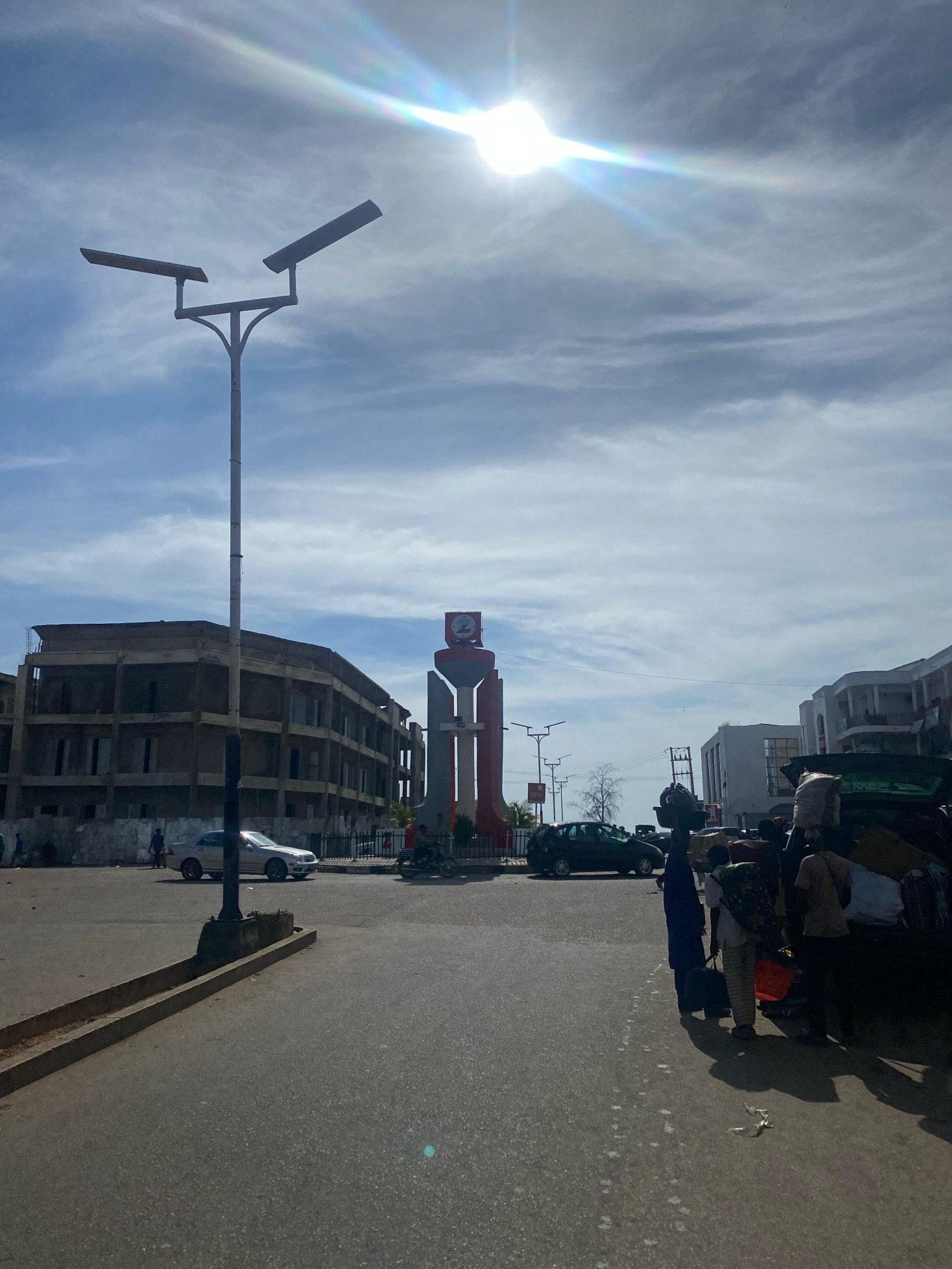 The image depicts a street scene in the city of Jos, Nigeria, likely near a roundabout or major intersection. In the foreground, a paved road stretches towards the background. To the left, a tall, modern streetlamp with two angled light fixtures stands prominently. Further back on the left, an unfinished or partially constructed multi-story building with exposed concrete and window frames dominates the skyline. A silver sedan is parked on the road in front of this building, and a few individuals are visible walking in the distance.
The center of the image features a large, red and white monument or sculpture, possibly a clock tower, with a distinctive shape. A traffic light is visible near the monument. The road continues past this monument, with more buildings and trees on the right side of the frame.
On the right side of the road, a group of people are gathered around a vehicle that appears to be heavily laden with luggage. The vehicle is dark-colored, possibly an SUV or van, with bags and other items piled on top and in the open trunk. Several individuals are standing or interacting near this vehicle, some carrying bags themselves. The sky above is bright blue with scattered white clouds, and the sun is high, creating lens flare and casting strong shadows on the ground. The overall impression is of a busy urban environment with ongoing activity, possibly related to travel or commerce.