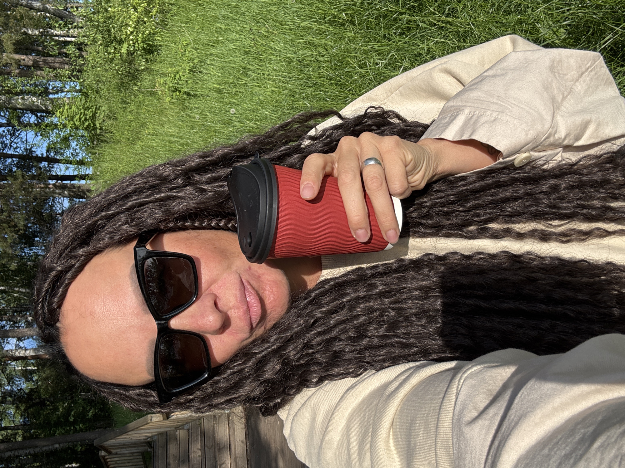 The image captures a selfie of a person relaxing outdoors in what appears to be Chaikovsky, Russia, on a bright, sunny day. The subject is seen from the chest up, with a significant portion of her long, dark, braided or twisted hair framing her face. She is wearing dark, square-framed sunglasses and a light beige or cream-colored long-sleeved top or jacket.
In her right hand, she holds a red, corrugated disposable coffee cup with a black lid. A silver-toned ring is visible on her left ring finger. The background features lush green grass, indicating a natural or park-like setting. Further back, slender trees with light-colored trunks (resembling birch) and green foliage are visible under a clear, bright blue sky. A wooden structure, possibly a deck or path, is partially visible in the bottom left of the frame. The overall impression is one of calm and enjoyment of the pleasant outdoor weather. No discernible text is visible on any objects.