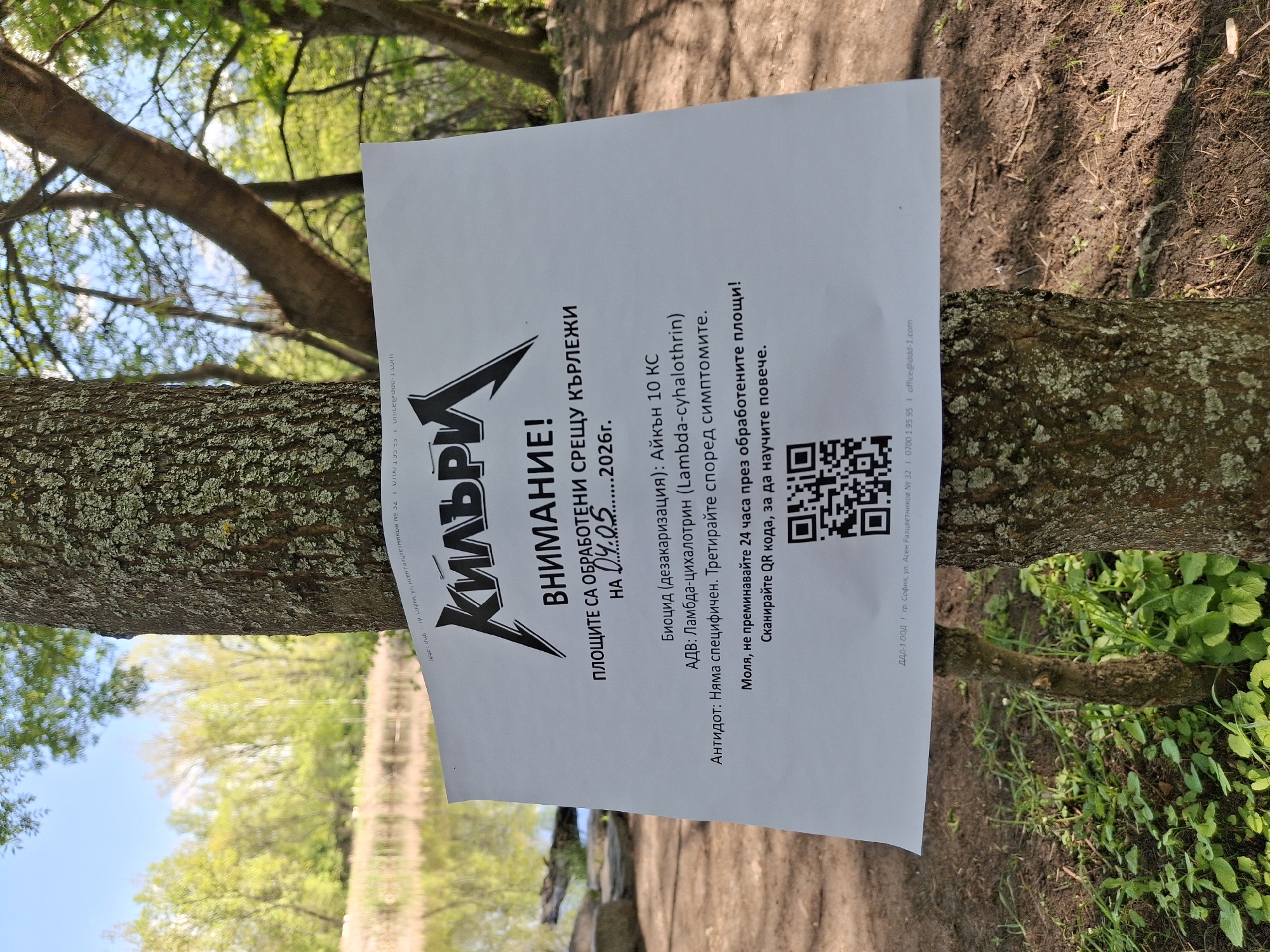A sign posted on a tree in an outdoor, park-like setting displays a warning in Bulgarian. The sign is titled "КИЛЪРИ" in large, stylized letters above the word "ВНИМАНИЕ!" (Attention!). It indicates that the area has been treated for ticks (КЪРЛЕЖИ) on 04.05...2026. The biocidal product used is Айкън 10 КС, with the active substance being Ламбда-цихалотрин (Lambda-cyhalothrin). It advises against crossing the treated areas for 24 hours and includes a QR code for more information. At the bottom, it lists "ДДЛ-1 ООД" from Sofia, at ул. Асен Разцветников № 32, along with a phone number and email address. The surroundings show a body of water, trees, and ground vegetation, suggesting a lakeside or wooded area in an urban park setting.