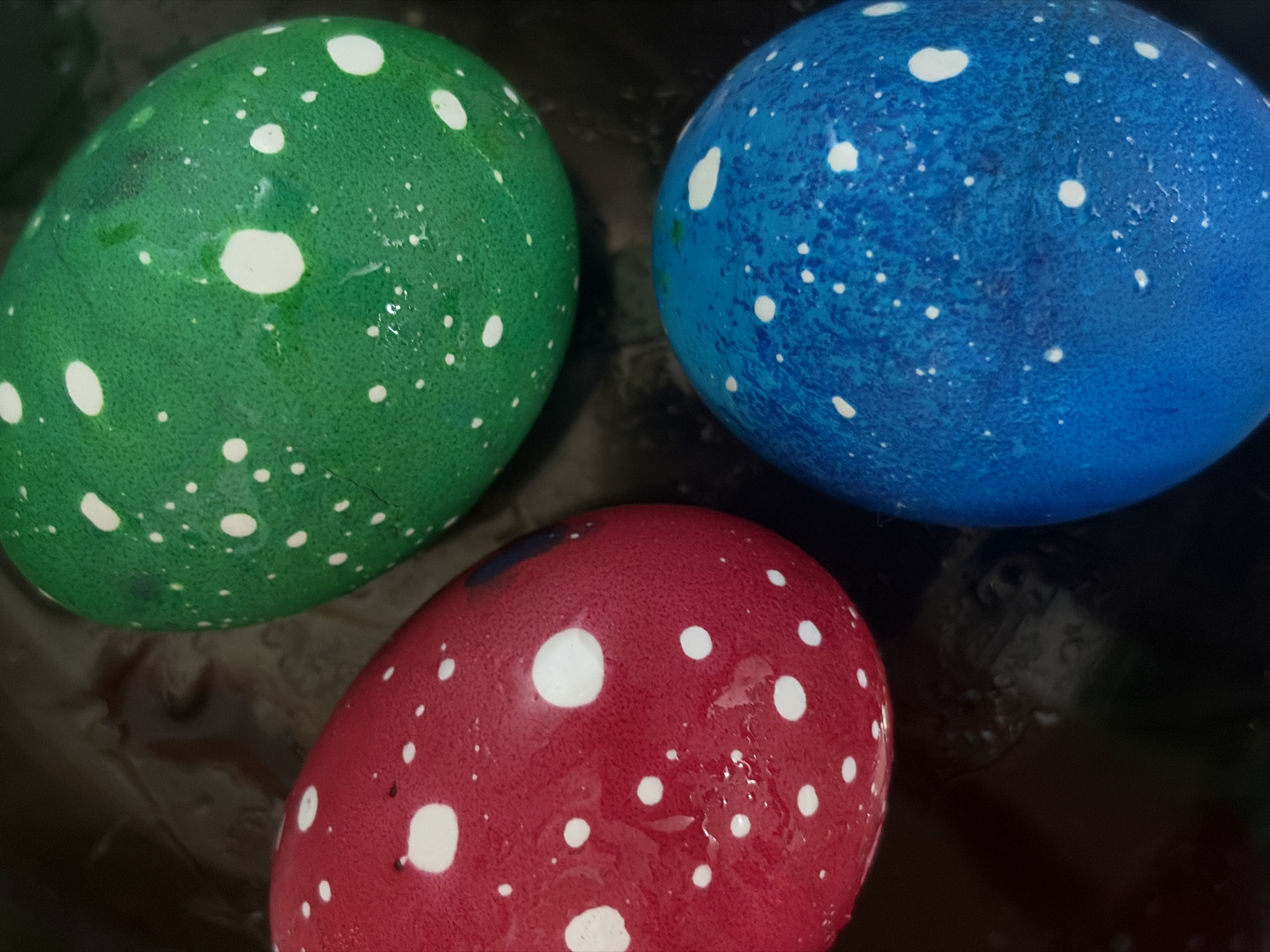 Three dyed Easter eggs are arranged in a close-up shot. The eggs are green, blue, and red, and all are speckled with white dots. The image is set against a dark, reflective background. There is no indication of location in Saint Petersburg, Russia, or any other geographical marker. The composition focuses solely on the eggs and their immediate surroundings.