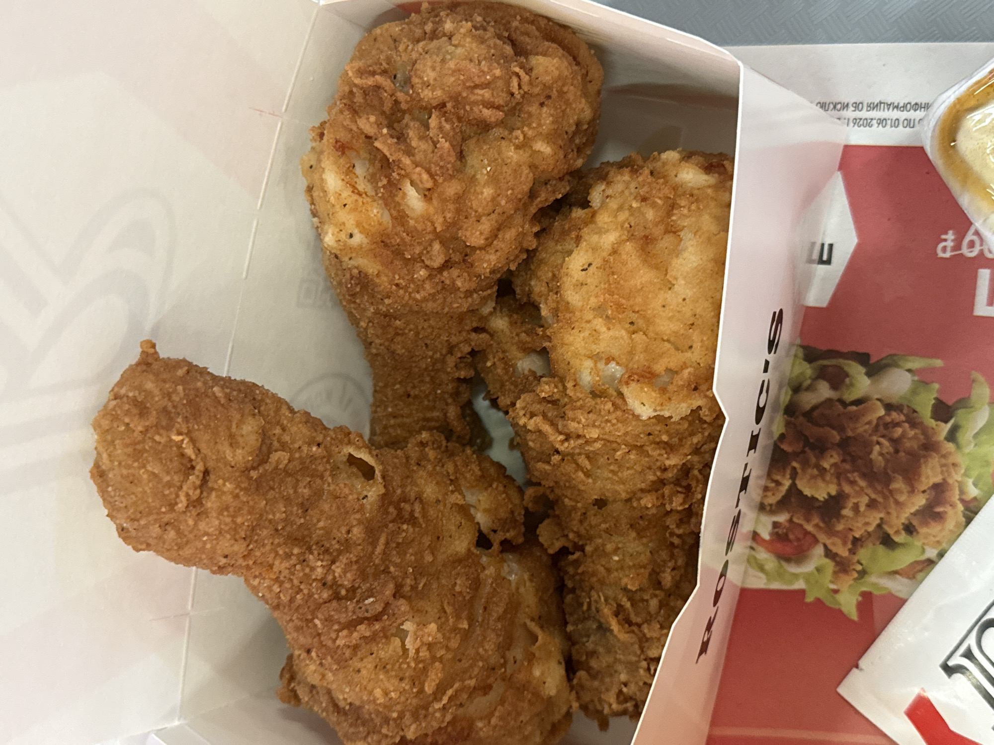 The image shows three pieces of fried chicken in a white and red KFC box. The location is Moscow, Russia, as evidenced by the Russian text on the box. The box also contains a packet of sauce and an image of a burger.