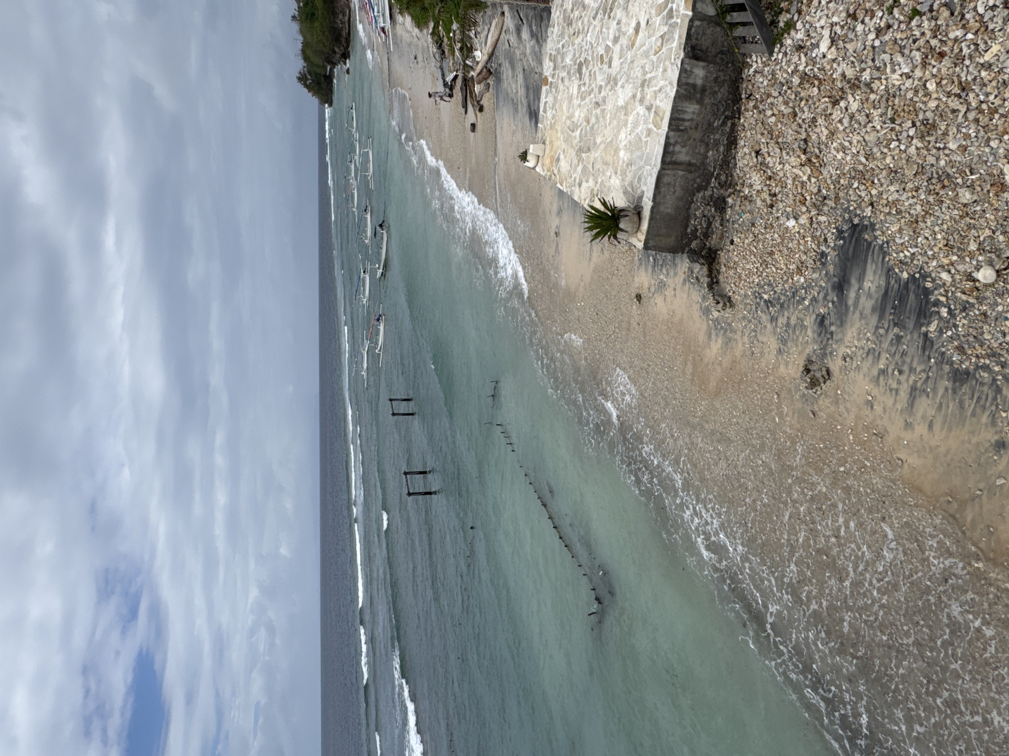 The image shows a scenic view of a beach in Nusa Penida, Indonesia. The ocean is a clear turquoise color with gentle waves rolling onto the shore. Numerous small, white, traditional Balinese boats are anchored in the shallow water. The beach itself is a mixture of sand and pebbles. In the foreground, there is a stone-tiled patio area with potted plants. Further up the beach, there are piles of driftwood and some wooden structures in the water. The sky is overcast with clouds. A single person is visible walking along the beach. The overall scene conveys a sense of tranquility and natural beauty.