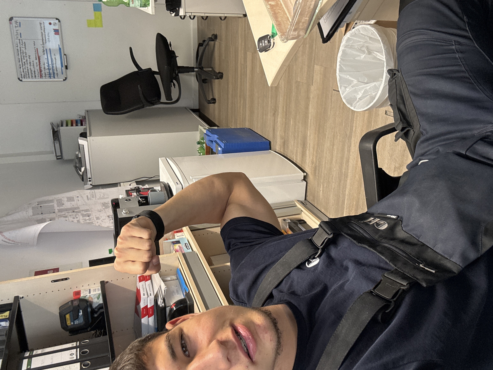 A male individual in a dark blue t-shirt and dark blue overalls is seated in an office chair, posing with his left arm flexed, displaying his bicep. He is looking towards the camera with his mouth slightly open. The background suggests an office or workshop environment. To the left, shelves are visible with binders and other office supplies. A whiteboard with writing is on the wall behind him. In the mid-ground, a white refrigerator and a blue storage container are present. In the foreground, the man's legs and the chair are visible, along with a white trash can. A desk with car keys and a container of food is to the right. There is no indication of the city of Mannheim, Germany, in the provided image.