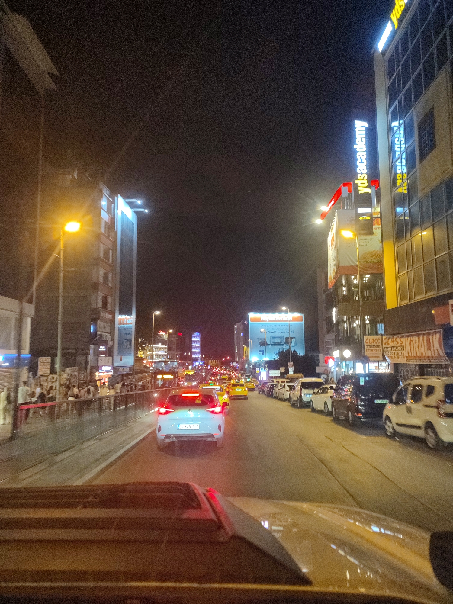 The image depicts a busy street scene at night in Kadıköy, Turkey. A multi-lane road is filled with traffic, primarily consisting of cars and yellow taxis. Pedestrians are visible on the sidewalk to the left, separated from the road by a metal railing. Buildings line the street, featuring illuminated signs, including a prominent vertical sign that reads "ydsacademy" and another large sign advertising "Hepsiburada." Streetlights cast a warm glow on the scene, and the headlights and taillights of the vehicles create streaks of light. The perspective suggests the photo was taken from inside a vehicle, looking forward. The overall environment is urban and active, characteristic of a commercial district at night.