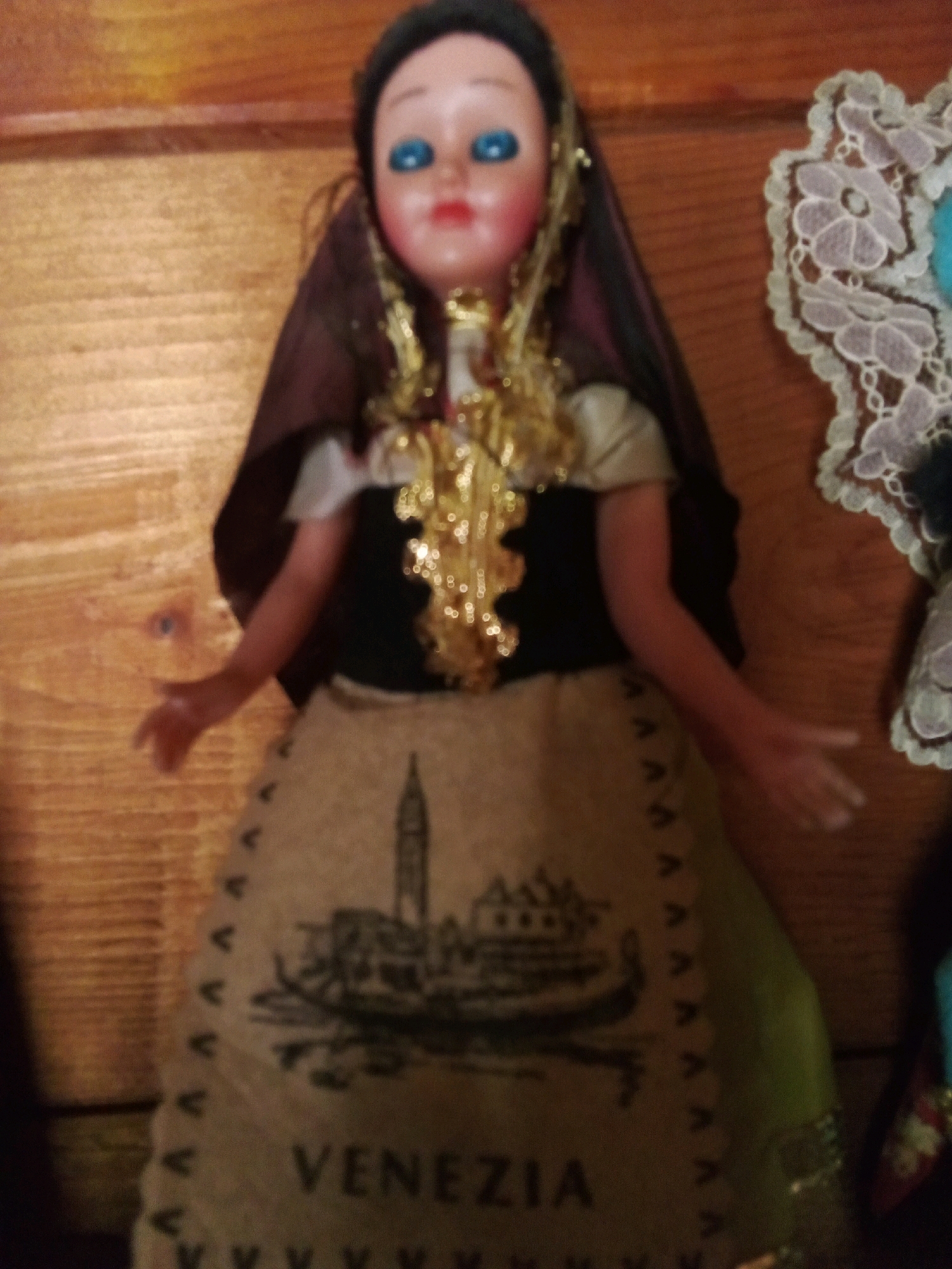 The image is a close-up, slightly out-of-focus shot of a doll dressed in what appears to be traditional Venetian attire. The doll has blue eyes and red lips. She wears a dark brown head covering, a black bodice, and a light brown skirt adorned with a silhouette of a gondola and buildings, along with the word "VENEZIA" printed in black. A decorative gold necklace hangs around the doll's neck. To the right of the doll, a piece of white lace trim is visible. The background is a wooden surface, likely a shelf or wall. There is no indication of the doll's location within the city of Santomenna, Italy, as the image does not contain any recognizable landmarks or contextual clues related to that specific place.