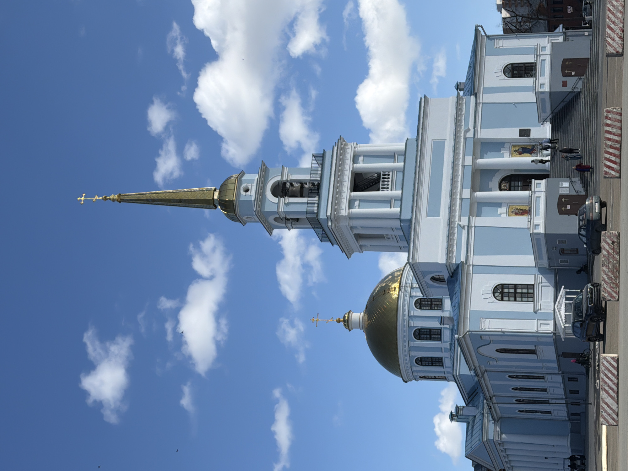 The image shows the Peter and Paul Cathedral in Votkinsk, Russia, angled upwards towards a blue sky with scattered white clouds. The cathedral's architecture features a prominent golden dome and a tall, slender spire with a golden cross at its apex. The building is painted in a light blue and white color scheme, and it includes multiple levels with windows and decorative columns. A few people are walking on steps to the side of the building. Vehicles are parked nearby, and striped barriers are present along the edge of the street.