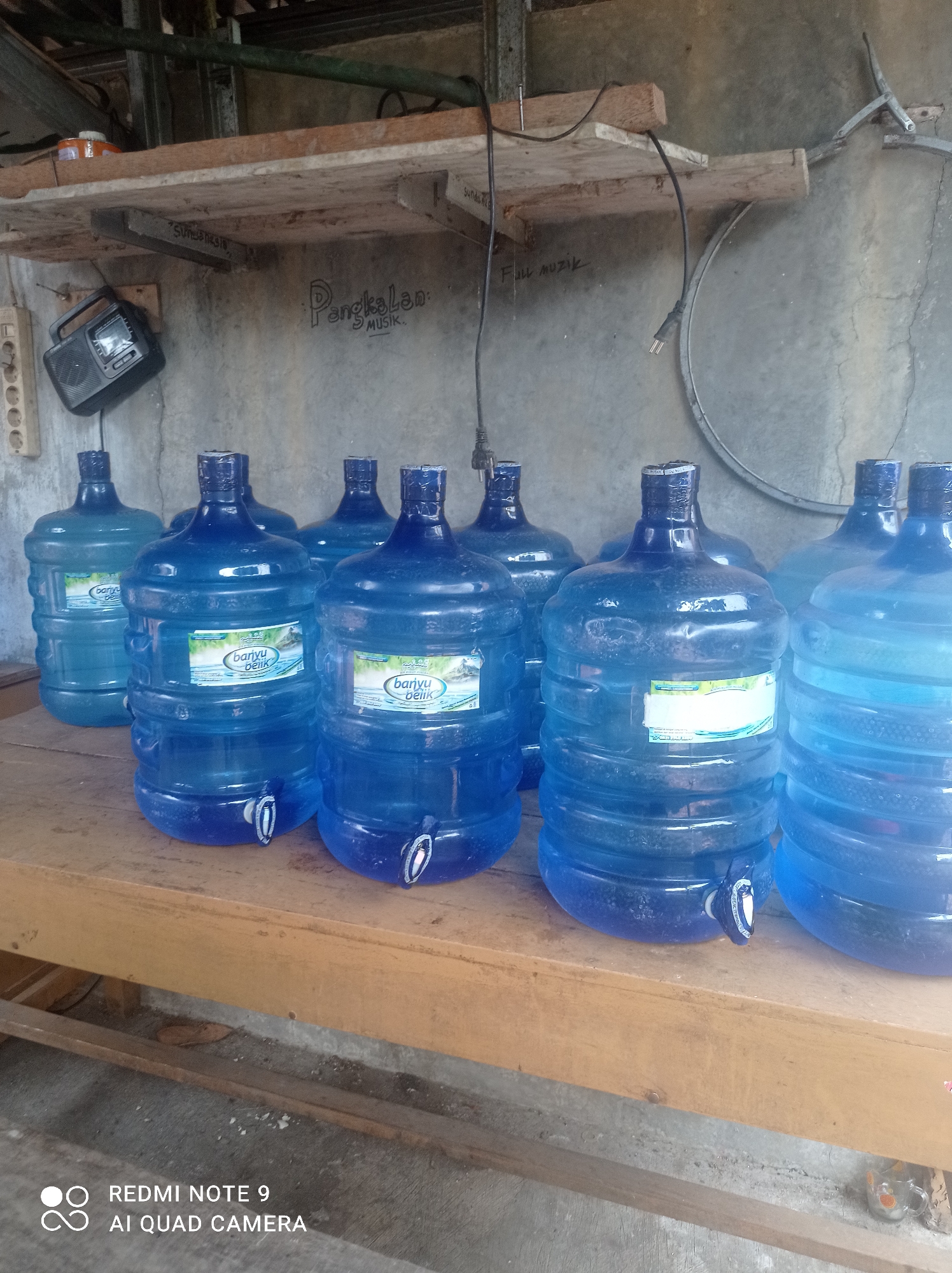 This image shows a collection of large blue plastic water jugs, many with spigots attached, arranged on a wooden surface. The jugs are filled with water and appear to be stored in an outdoor or semi-outdoor environment. In the background, a concrete wall is visible, with some handwritten text on it. A shelf is mounted above the wall, holding a small radio and electrical components. The overall impression is of a functional space, possibly a storage area or a point of sale for bottled water. The presence of the text "REDMI NOTE 9 AI QUAD CAMERA" in the bottom left corner indicates that the image was taken with a Xiaomi Redmi Note 9 smartphone. The text "PangkalAn : MUSIK." is visible on the wall, which translates to "Place : MUSIC." There is also text that reads "Full muzik" on the wall.