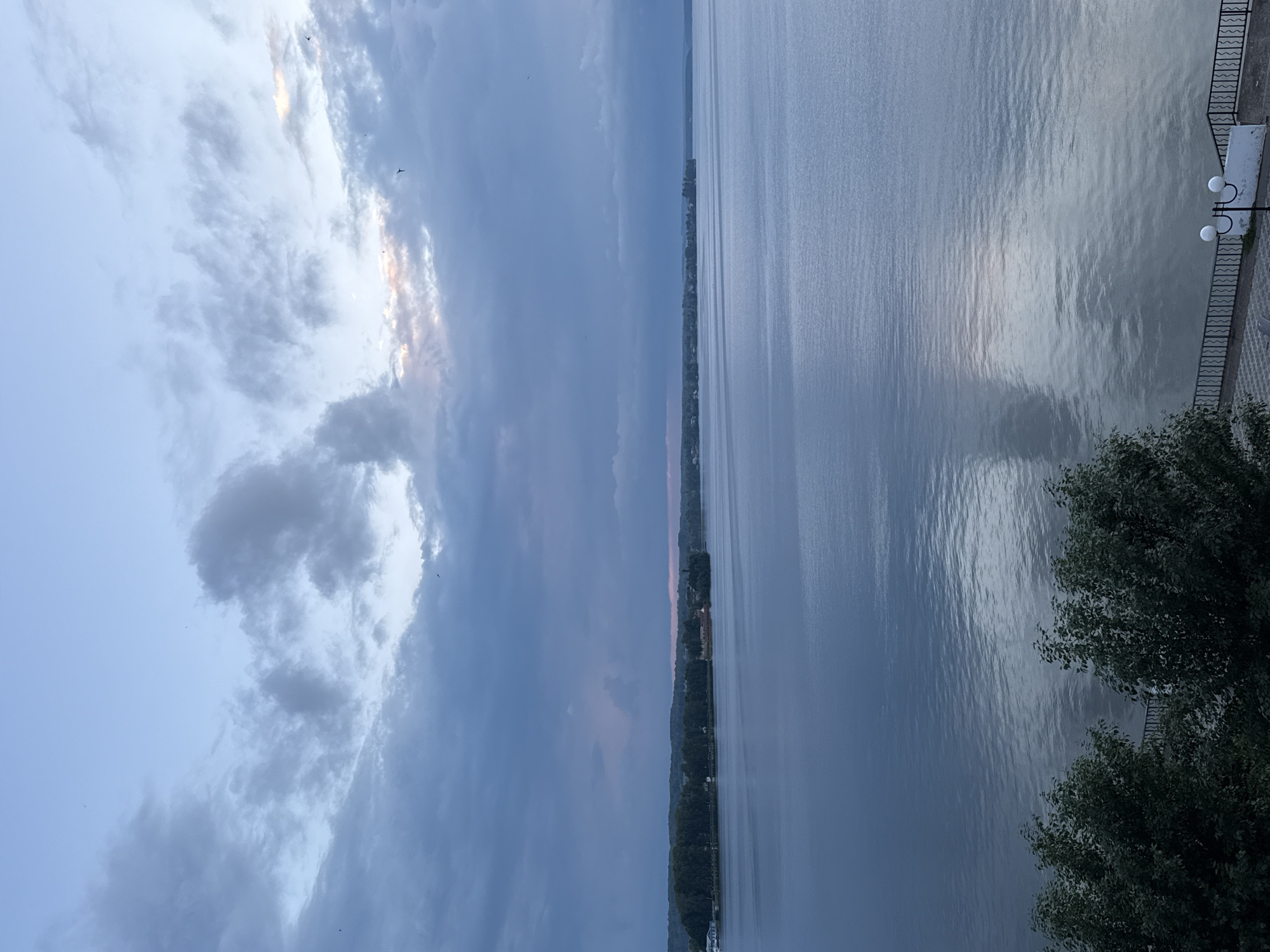 A body of water, likely a lake or river, stretches across the frame under a cloudy sky. The water is a calm, muted blue, reflecting the sky and clouds. In the distance, a tree-lined shore is visible, with hints of buildings and structures suggesting a populated area. The foreground features dark green foliage from trees, obscuring the lower part of the scene. A decorative railing and a lamppost are partially visible at the bottom right, indicating a walkway or promenade. The overall atmosphere is serene and tranquil, with the soft light suggesting either early morning or late evening. The image is identified as being taken in the city of Votkinsk, Russia.