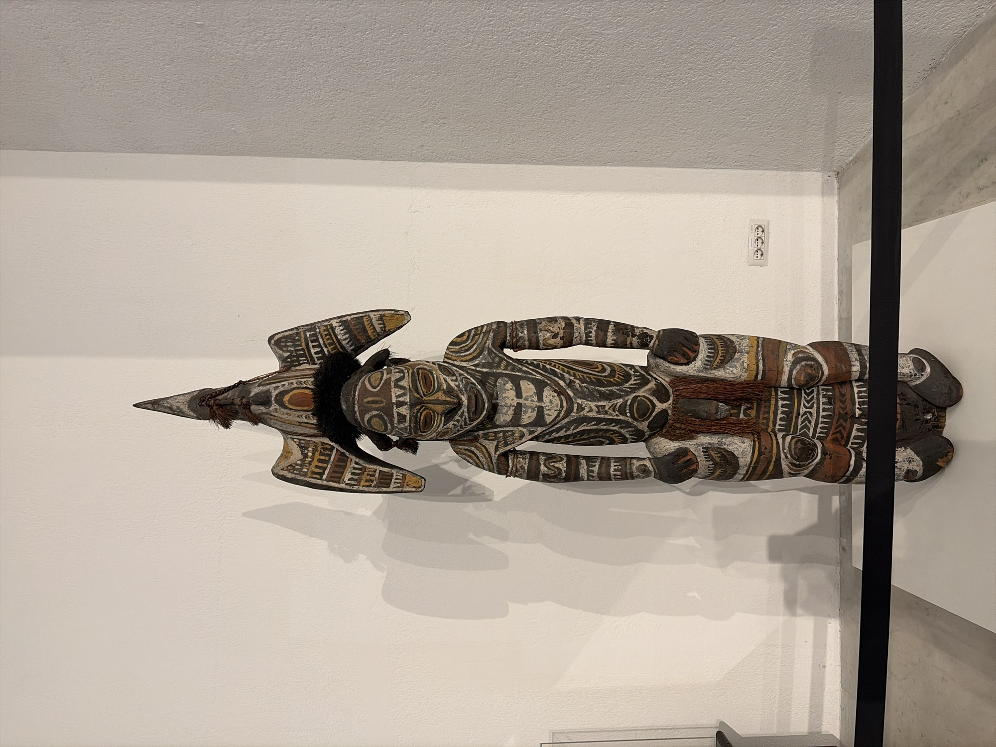 An elaborately carved and painted wooden effigy stands against a white wall in a museum in Vatican City. The effigy depicts a figure with a stylized human face, adorned with black hair and a headdress resembling a bird with outstretched wings. The body is decorated with intricate geometric patterns in white, brown, and yellow. The figure is positioned in a squatting posture, with its hands resting on its knees. A dark barrier tape runs horizontally in front of the effigy. The background shows a white wall with subtle shadows cast by the effigy. A triple electrical socket is visible on the wall to the right. The flooring appears to be a light-colored marble.