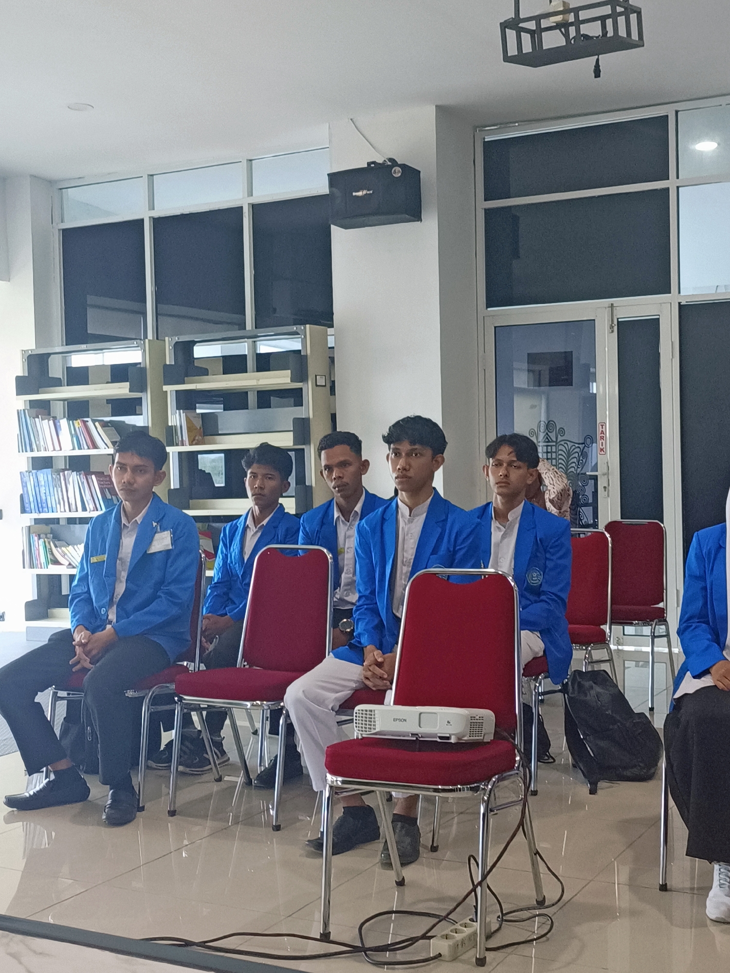 In a brightly lit indoor space resembling a library or classroom, a group of five young men, dressed in matching blue blazers over white shirts, are seated on red chairs. The men appear to be students, attentively facing forward. In the foreground, a white Epson projector rests on a red chair. Electrical cords and a power strip are visible on the tiled floor. Behind the seated individuals, rows of bookshelves filled with books extend along the wall, suggesting an academic or educational setting. Large windows with blinds are present in the background. The overall atmosphere is one of a formal gathering or presentation. The location is identified as Mesjid Reuntoh, Indonesia.
