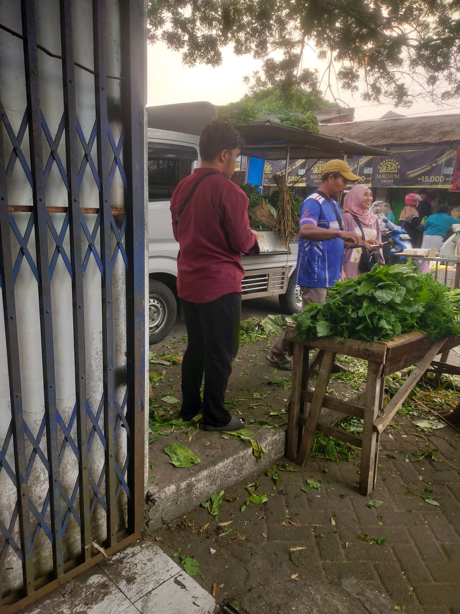 The image depicts an outdoor market scene in Bululawang, Indonesia. In the foreground, a metal accordion gate stands on the left, partially obscuring the view. To the right of the gate, a man in a maroon shirt and black pants stands with his back to the camera, facing a market stall.
The market stall features a wooden table laden with a large pile of fresh green leafy vegetables, likely for sale. Behind the table, a man wearing a blue shirt with a white stripe and a yellow cap is interacting with a woman in a pink headscarf. They appear to be engaged in a transaction or conversation. Further behind them, a white truck is visible, and other market-goers and stalls are present in the background, suggesting a busy commercial area. The ground is paved with dark bricks, and scattered leaves and vegetable trimmings are visible around the stall, indicating typical market activity. The overall lighting suggests it might be early morning or late afternoon, with diffused sunlight filtering through tree branches overhead.