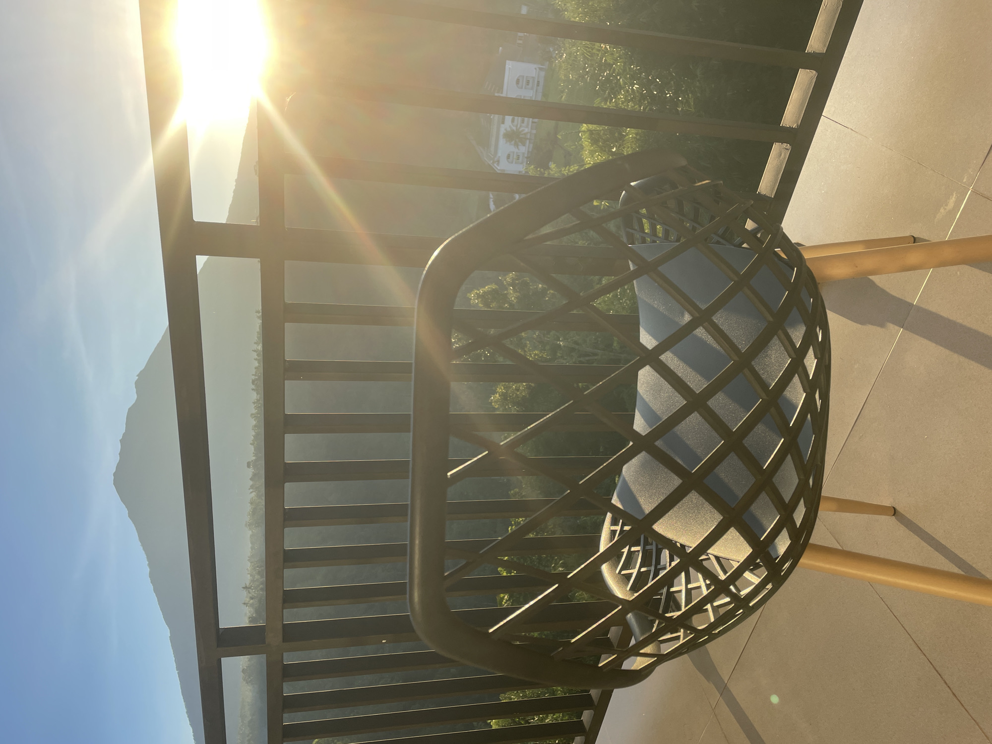 A medium shot of a black, modern chair with a gray cushion sitting on a balcony. The balcony overlooks a lush green landscape with a mountain peak visible in the background and is likely taken in Rendang, Indonesia. The sun is high in the sky, causing a bright glare. The balcony has a dark metal railing. The floor of the balcony is tiled.