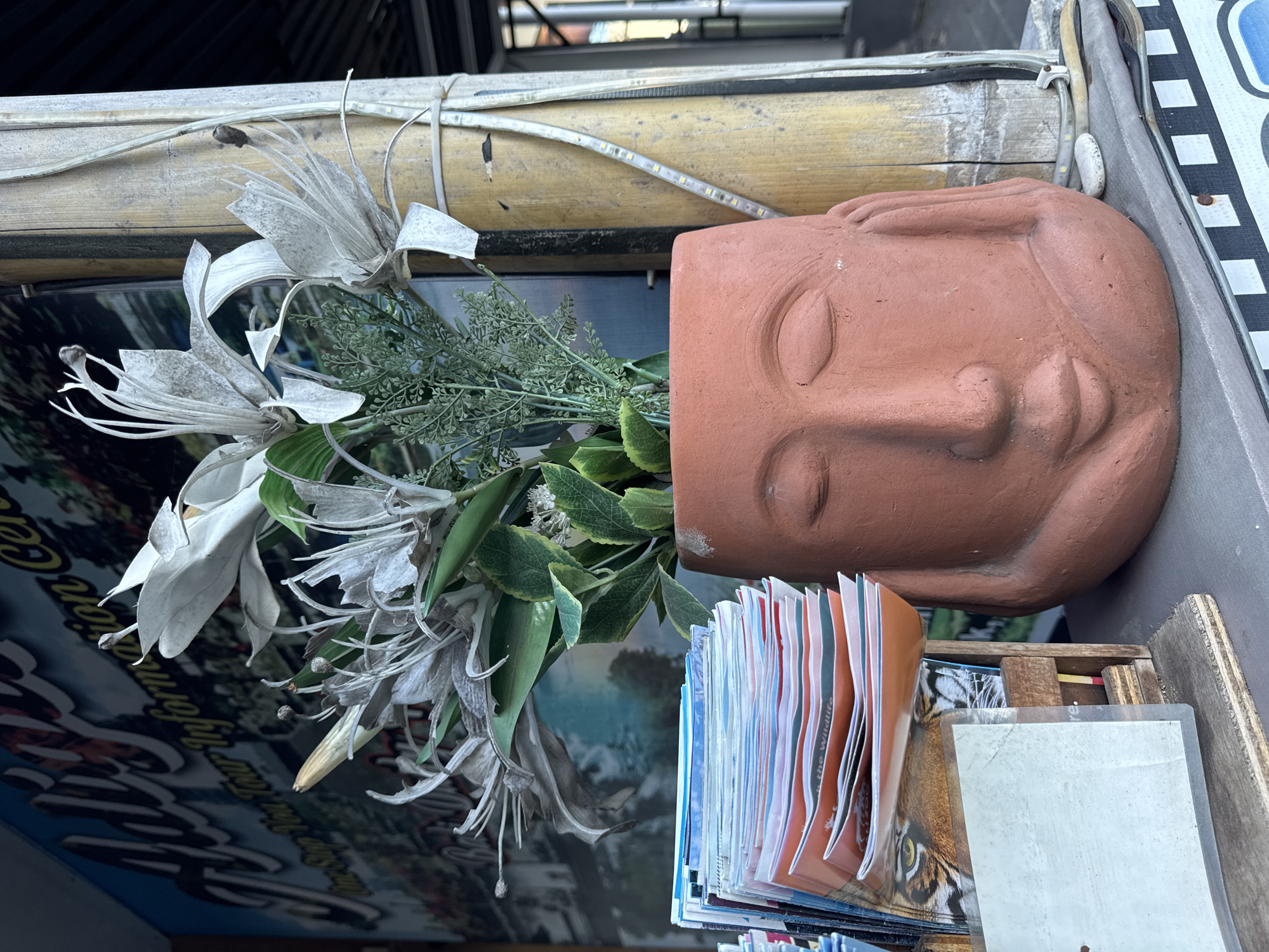 A terracotta head-shaped planter is positioned on an elevated surface. Dried flowers and green foliage sprout from the top of the planter, which rests near a stack of magazines and a wooden container. A bamboo pole is strapped to the structure behind the planter, and an LED strip is attached to the pole. A sign or poster is visible in the background. The location is Kuta, Indonesia.