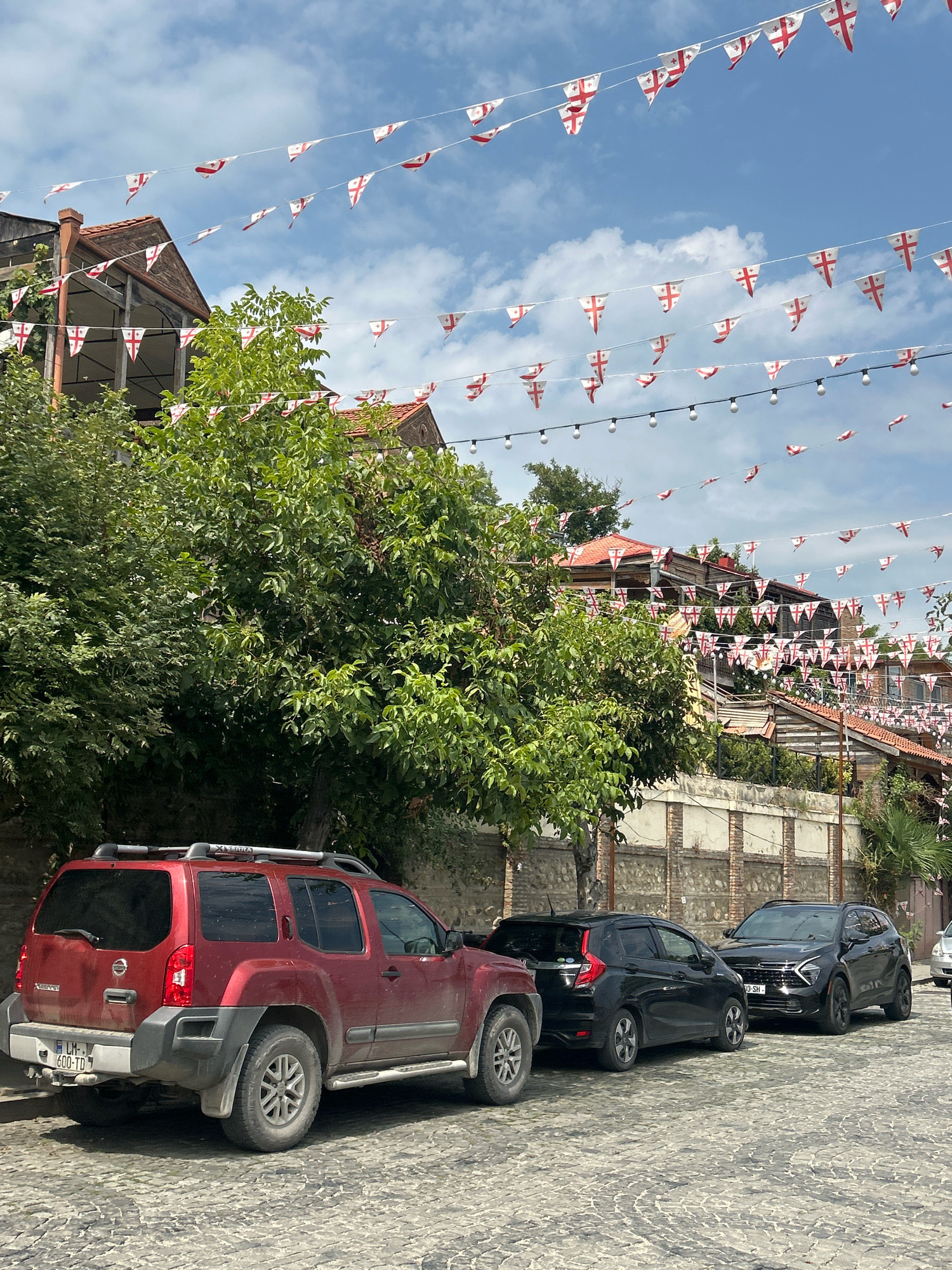The image shows a cobblestone street in Sighnaghi, Georgia, lined with buildings adorned with strings of Georgian flags. In the foreground, three parked cars are visible: a red SUV on the left, followed by two black cars. Lush green trees partially obscure the buildings, which feature traditional architecture with reddish-brown roofs. The sky is blue with scattered clouds. The scene depicts a street scene in a Georgian town, likely during a festive occasion or celebration due to the abundant display of national flags.