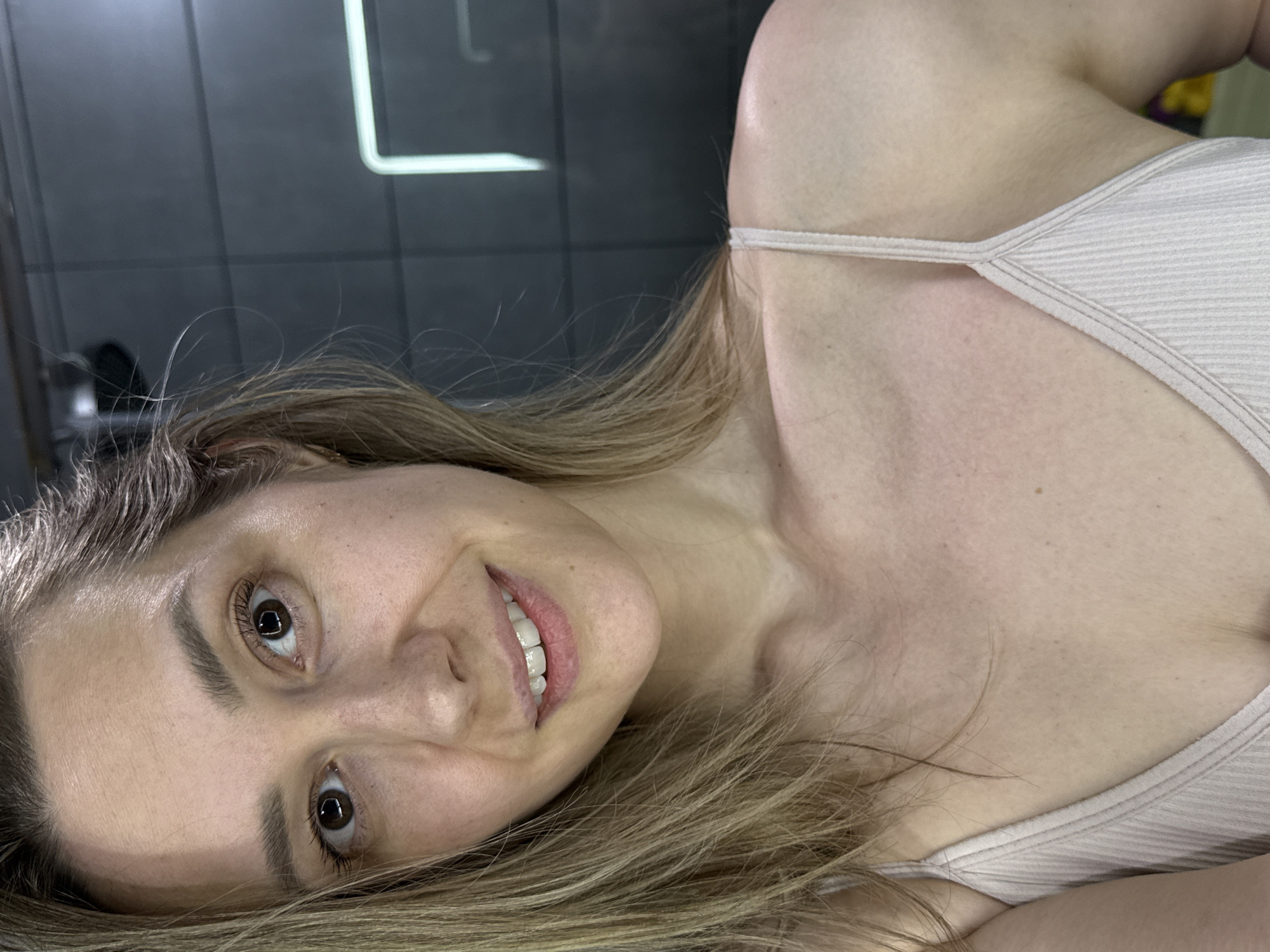 A woman is lying on her side, smiling, facing the camera. She has light brown hair, brown eyes, and fair skin. She is wearing a light-colored ribbed tank top. The background is a dark, smooth surface with a rectangular light fixture visible. There is no information available to confirm this image was taken in the city of Chaikovsky, Russia.
