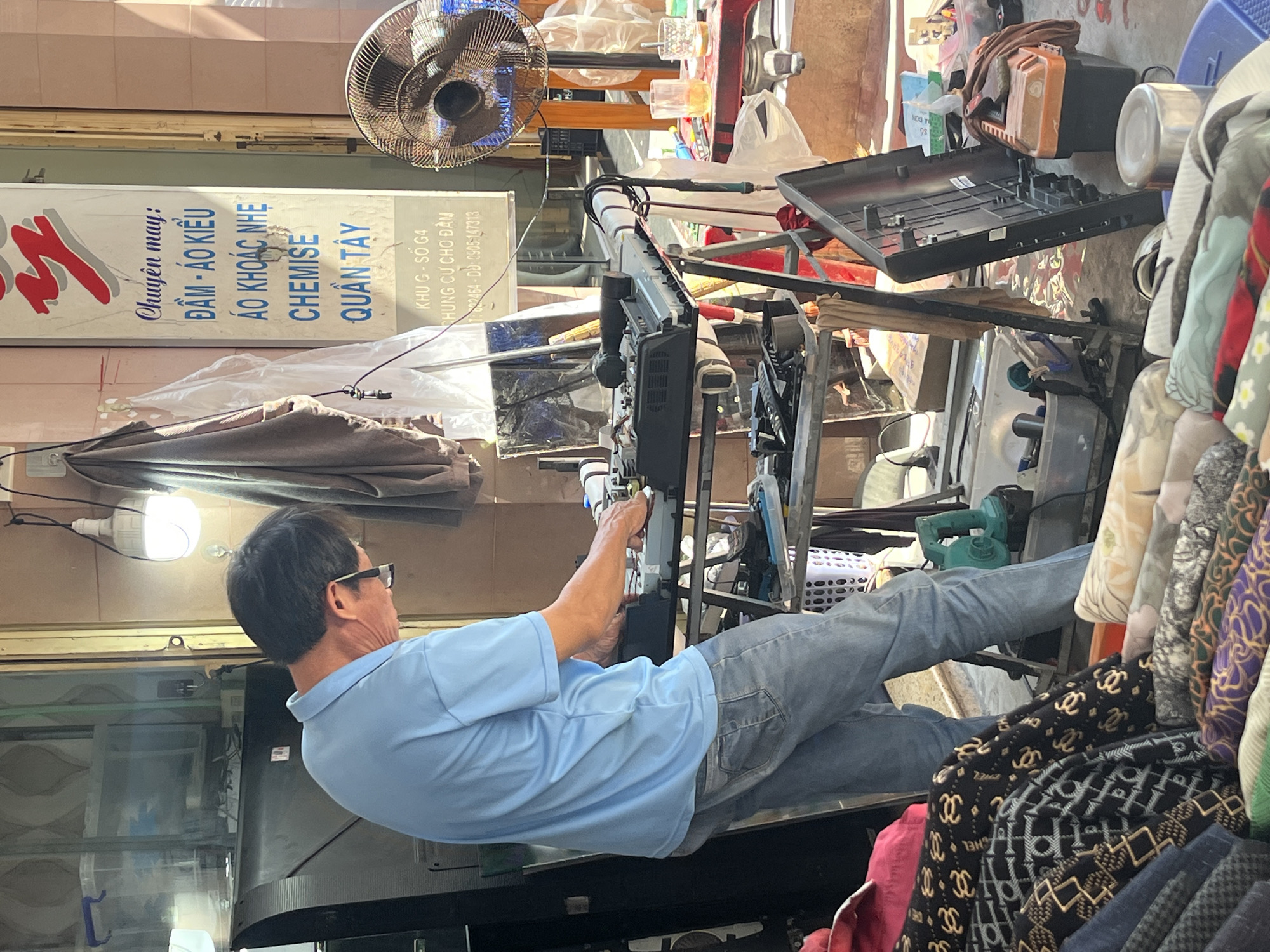 The image captures an interior scene in Nha Trang Ward, Vietnam, where a man is shown working on what appears to be a complex machine. He is wearing a light blue shirt and blue jeans, and glasses. The background is cluttered with various objects, including a fan, a sign in Vietnamese advertising clothing services (dresses, coats, shirts, and pants), toolboxes, textiles, and other materials. A light bulb illuminates the scene.