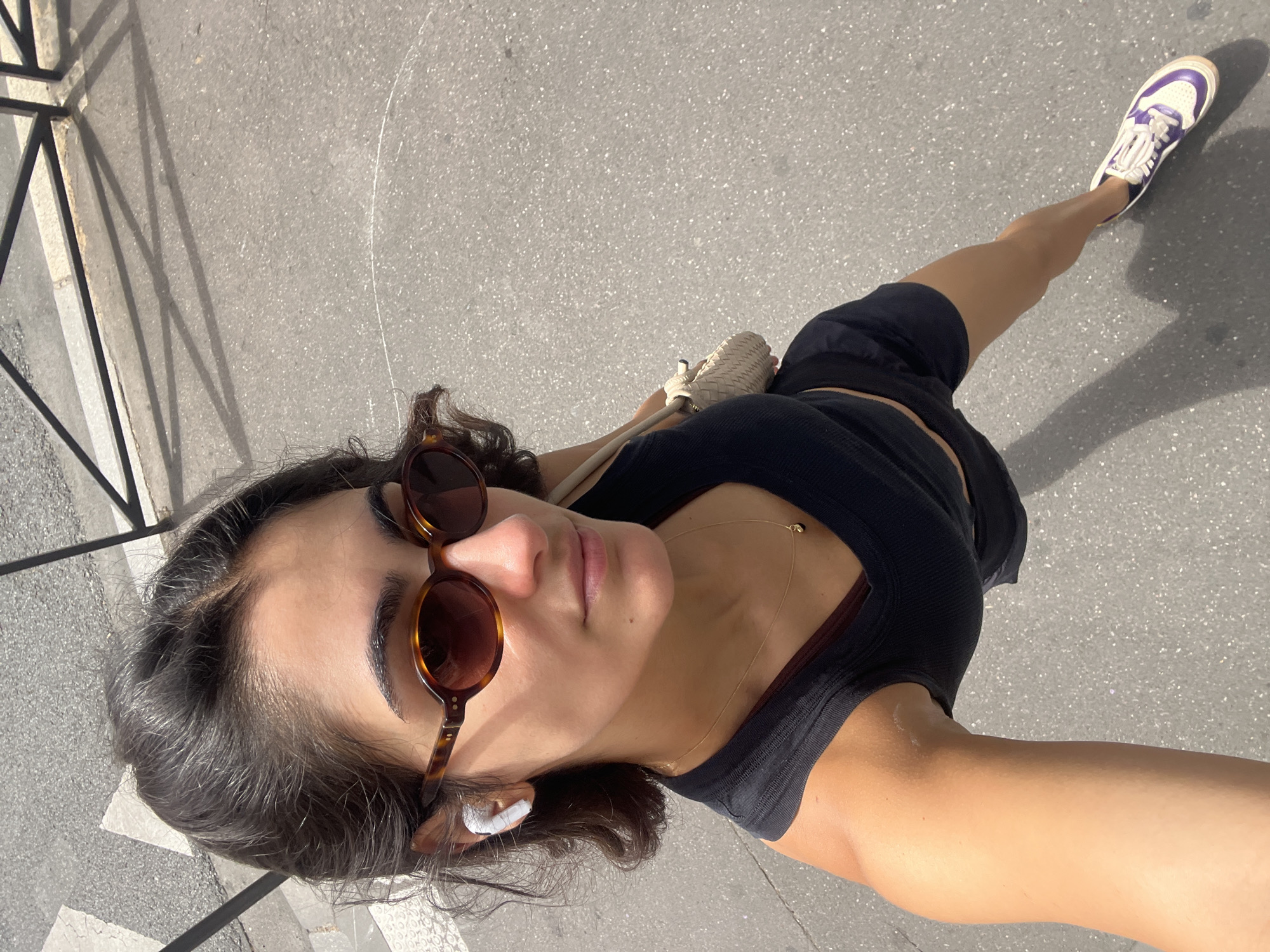 A woman is walking on a street in Paris, France, viewed from a high angle. She has dark hair, is wearing sunglasses, a black tank top, black shorts, and white and purple sneakers. She has a small woven bag on her shoulder and is wearing white earbuds. The street is paved and there is a metal fence and shadows on the ground.