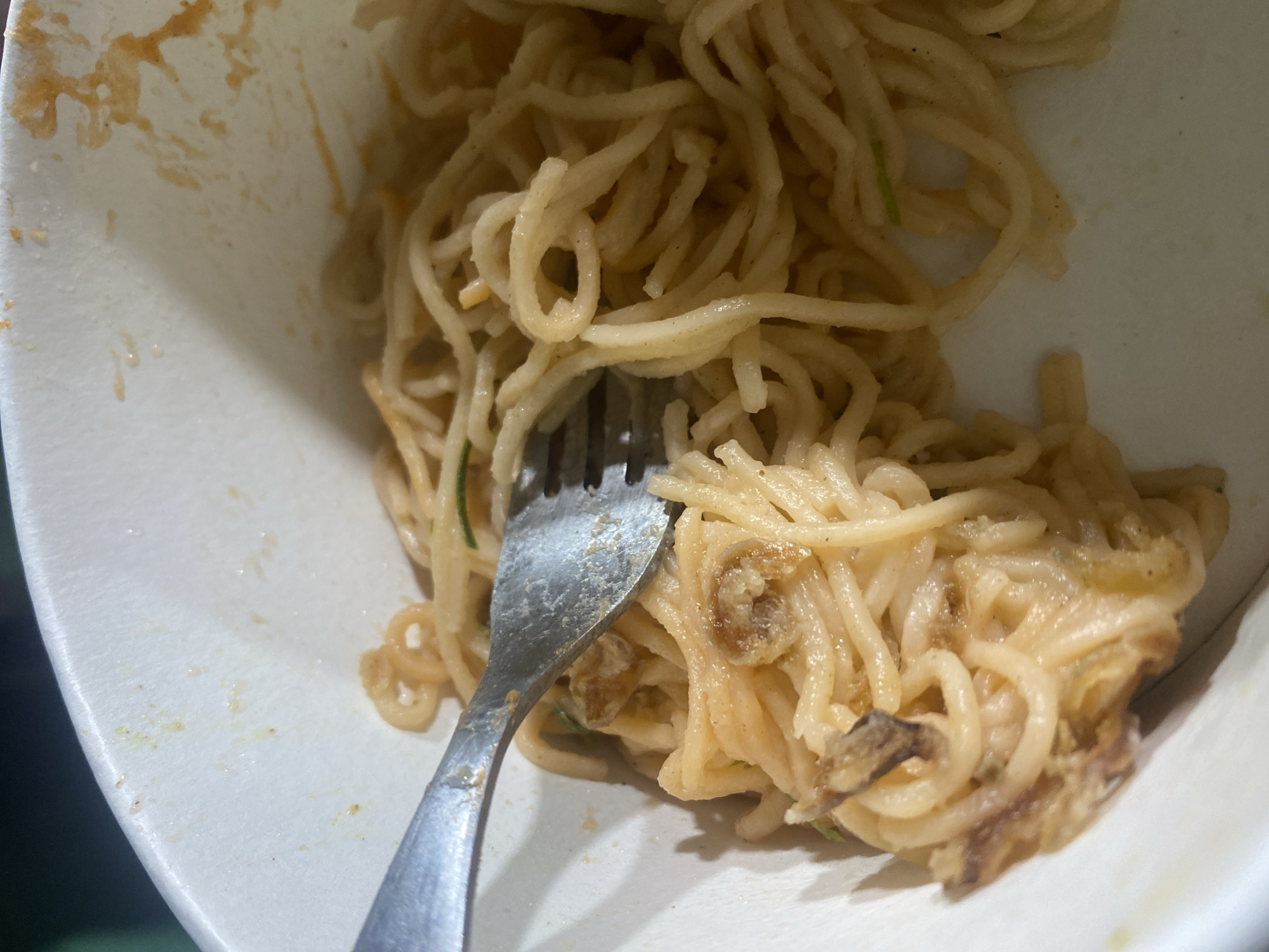 A close-up, overhead shot shows a white bowl containing a portion of spaghetti with a creamy sauce and some pieces of what appear to be mushrooms or meat. A silver fork is embedded in the pasta on the left side of the bowl. The rim of the bowl shows some slight staining from the food. The lighting is dim, casting shadows on the pasta. This scene is located in Kasihan, Indonesia.