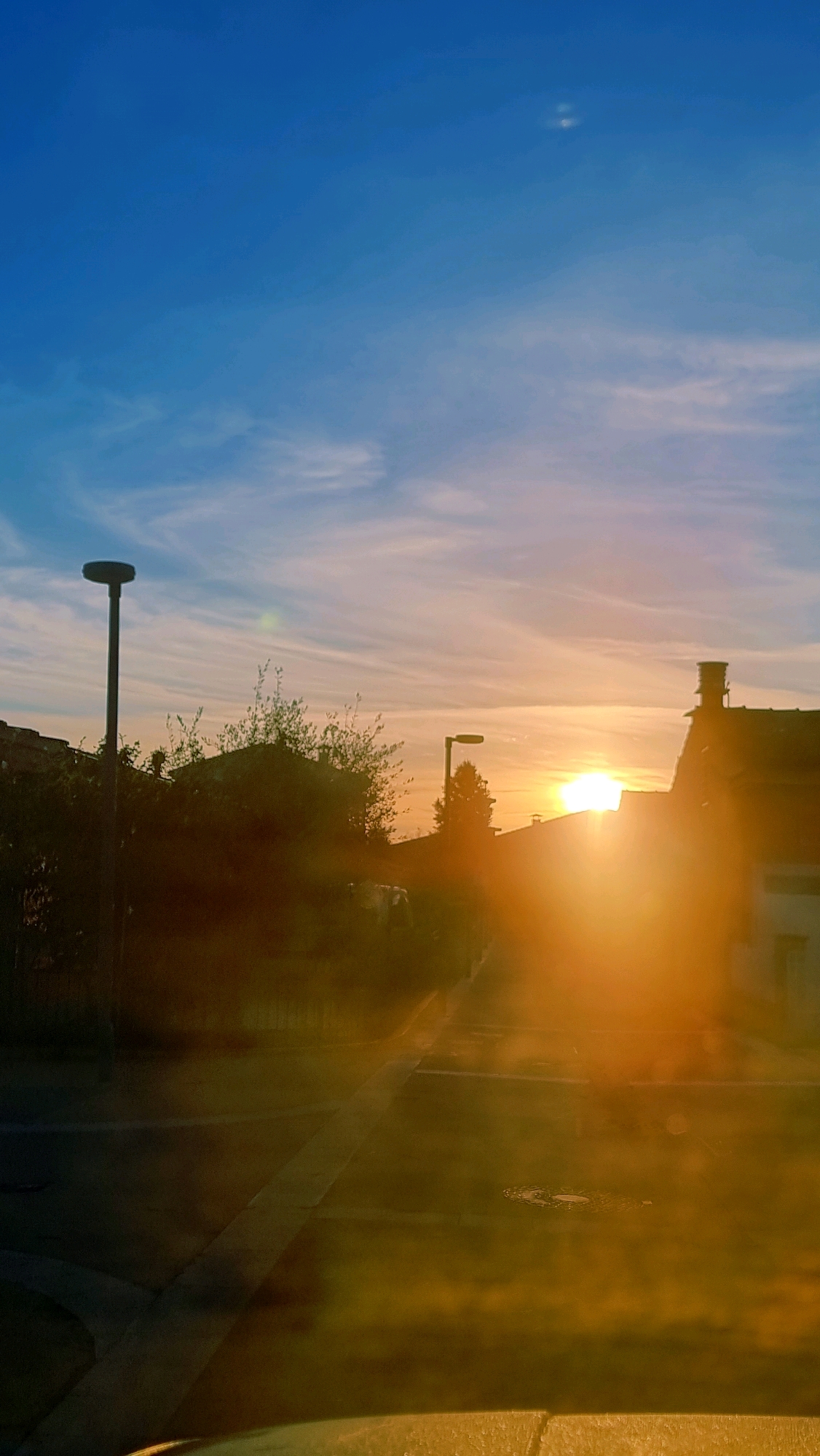 A vertical, medium-shot, exterior, eye-level, daytime photograph shows a street scene in Sant Joan Les Fonts, Spain, during a sunset. The sky is a gradient of deep blue at the top, fading to a light blue with wispy white clouds near the horizon. The sun, a bright orb of light, is low on the horizon, casting a warm, orange glow across the scene.
In the foreground, a street is visible, with a faint white line marking the center. The street appears wet, possibly from recent rain, reflecting the sunset's light. On the left side of the street, a tall, dark lamppost stands erect. Behind the lamppost, a cluster of green trees and some houses with dark roofs are partially obscured by the low-lying sunlight.
Further down the street, another lamppost is visible, also dark. To the right of this lamppost, the silhouette of a house with a chimney is prominent. The house also has a dark roof. The sunlight is so intense in this area that it creates a bright glare, obscuring finer details.
The overall atmosphere is serene, with the sunset casting a tranquil mood over the suburban landscape. There are no people visible in the frame.