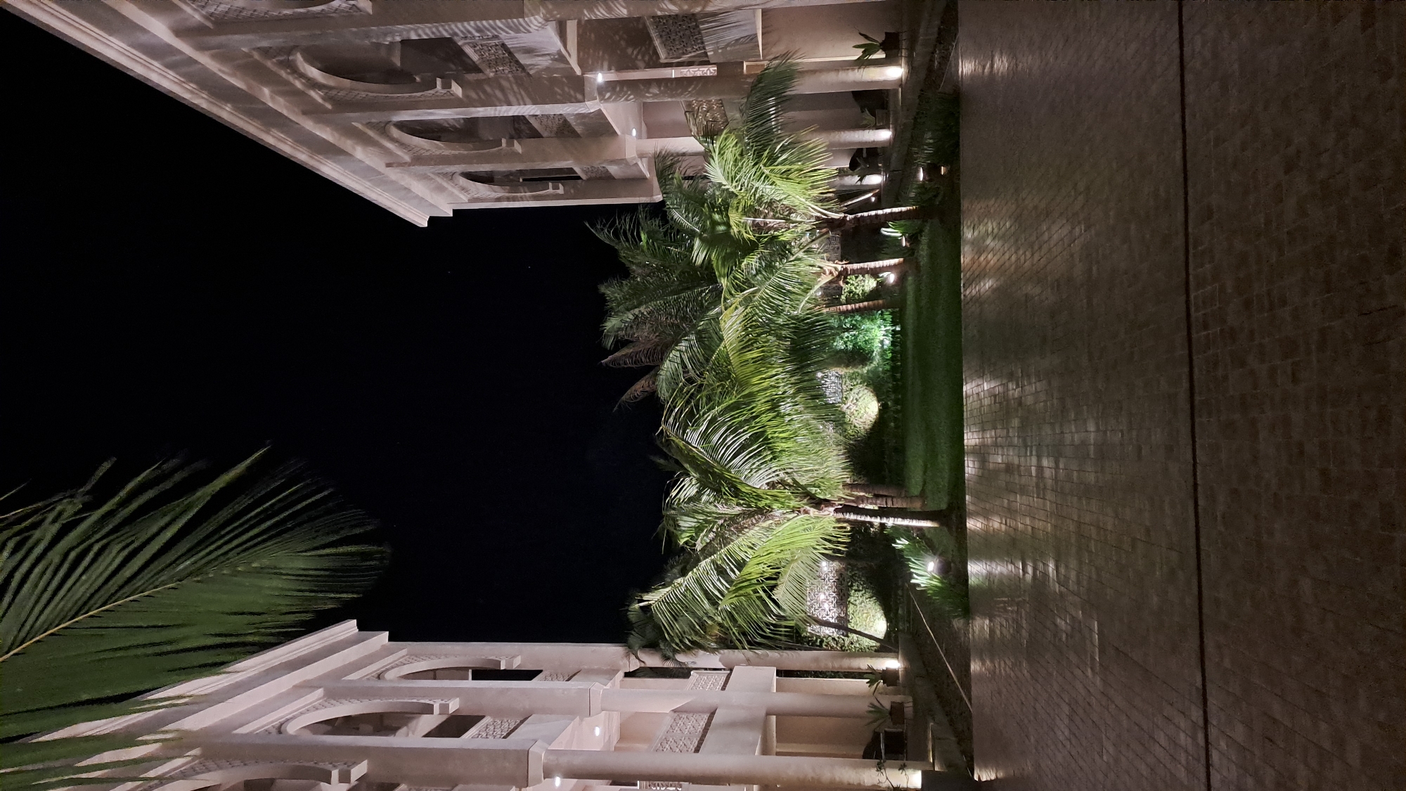 This image depicts an outdoor courtyard at night, likely within a resort or hotel complex in Daroykameu, Indonesia. The scene is illuminated by artificial lighting, creating a warm glow on the paved courtyard and the surrounding vegetation.
On either side of the courtyard are two identical buildings, characterized by their light-colored facades and arched openings. These buildings suggest an architectural style that might be influenced by traditional Islamic or Moroccan design. Palm trees with lush green fronds are scattered throughout the courtyard, some of them silhouetted against the dark night sky. The ground in the foreground is paved with rectangular stones, reflecting the ambient light.
The overall atmosphere is serene and tranquil, suggesting a peaceful nighttime setting. There are no people or animals visible in the frame. The focus is on the architecture, landscaping, and the interplay of light and shadow.