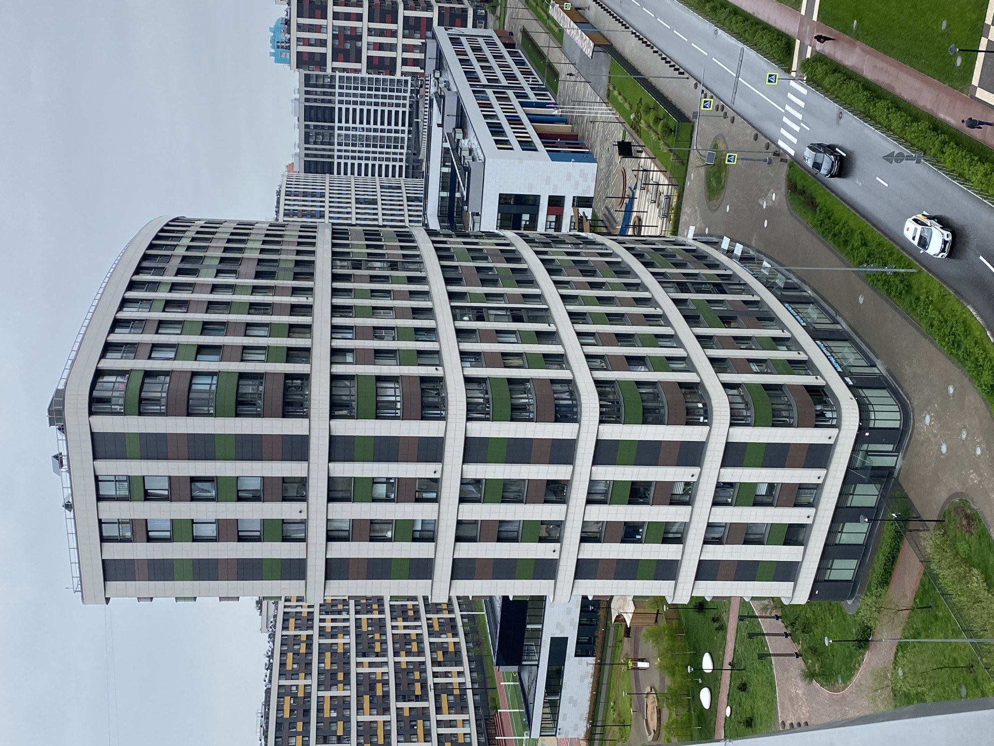 The image presents an elevated view of a modern urban landscape, likely within a residential complex. The location is in an unknown part of Russia. The focal point is a multi-story, curved apartment building, clad in a grid of windows and alternating horizontal stripes of light gray, green, and brown. Other apartment buildings of varying styles are visible in the background, some with white exteriors and colored accents, others with yellow and brown patterns.
A road with two cars is present, one white, one dark gray. Along the roadside, there are sidewalks, grass areas, and pedestrian crossings. There are street lights. The environment suggests a recently developed or rapidly growing area.