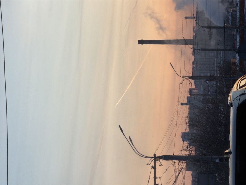 A sideways view shows a cityscape under a dusky sky. A jet contrail cuts diagonally across the sky. Chimneys emitting smoke rise from industrial buildings. Utility poles with streetlights and a dense network of wires dominate the foreground. The side of a car is visible on the right. The overall scene evokes a sense of urban activity blending with atmospheric phenomena.