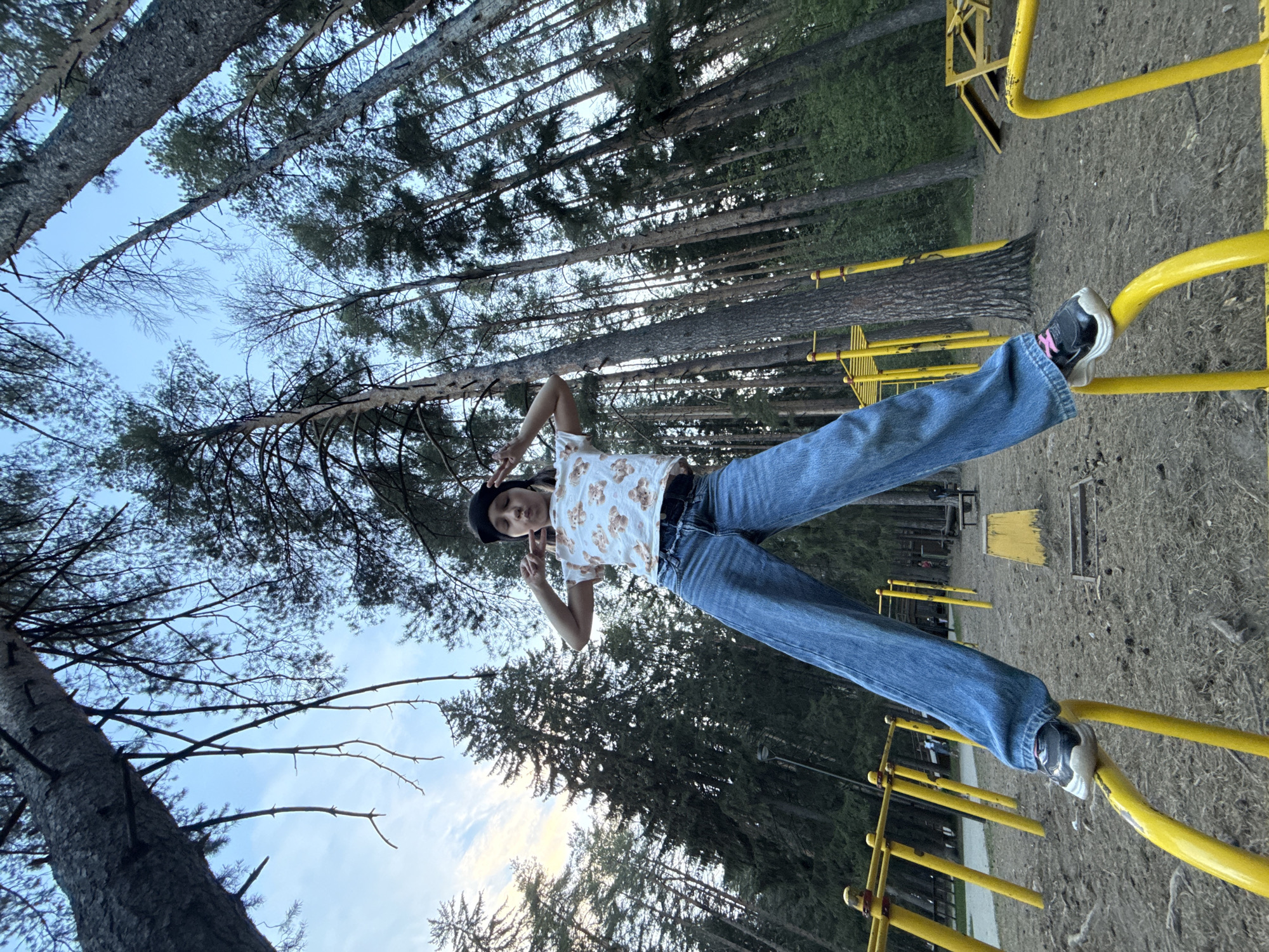 A person wearing a white shirt with a bear pattern, blue jeans, and a black hat is perched atop a yellow metal structure in a wooded area. The person is positioned horizontally, with their feet on the right side of the frame and their head on the left. The individual's hands are raised near their head, forming a "peace" sign with their fingers. The background consists of tall trees reaching towards a blue sky with some visible clouds. The ground is covered with dirt and sparse vegetation. The location is likely a park or recreational area in the city of Votkinsk, Russia, suggested by the presence of the park-like setting and the metal structure resembling playground equipment.