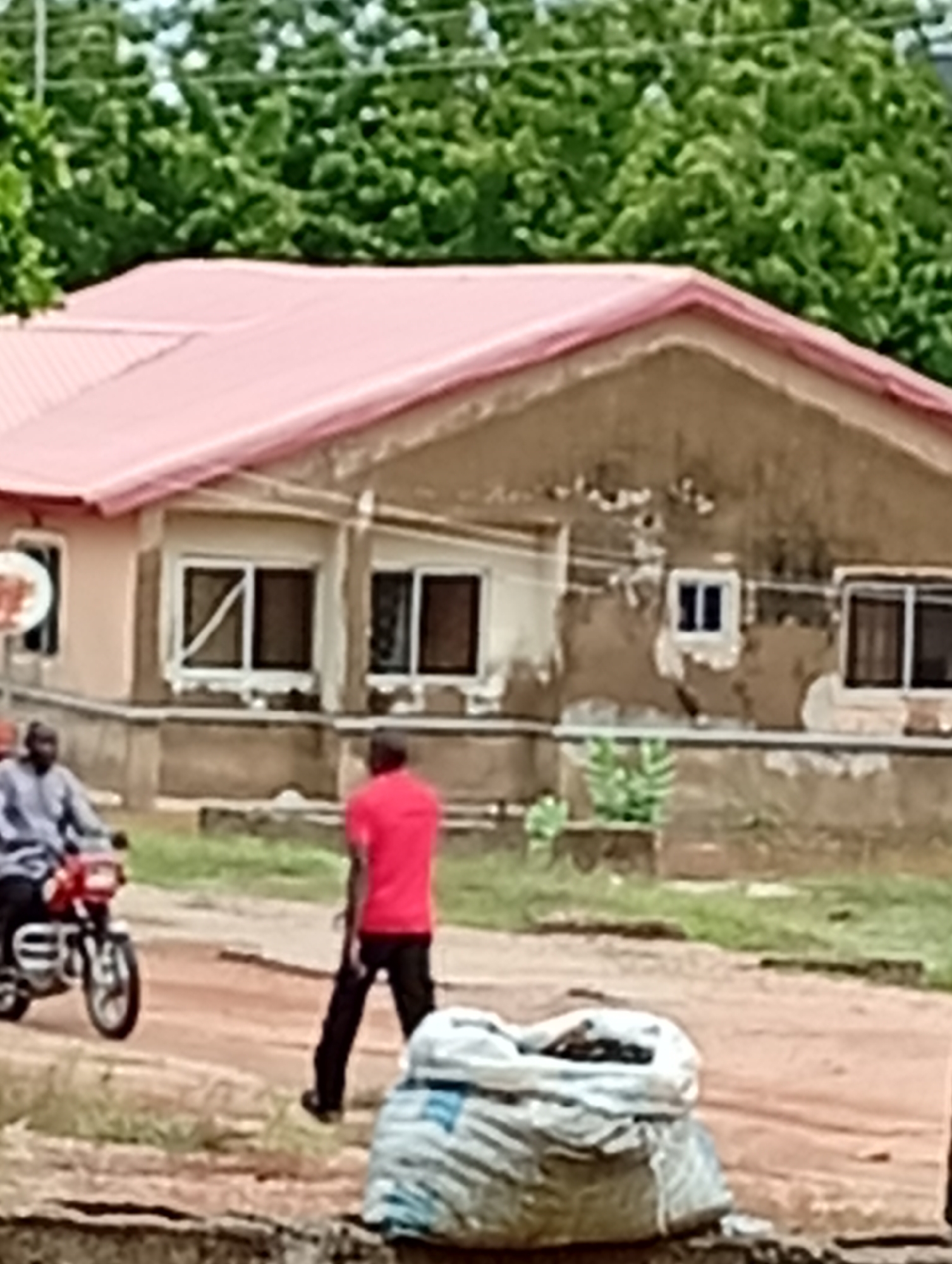The image shows a single-story building with a pink roof and peeling, tan-colored walls. There are several windows with dark frames. In front of the building is a dirt road. A man in a red shirt is walking on the road, and a man on a red motorcycle is driving in the opposite direction. A large bag sits in the foreground. The image appears to be taken outdoors in Unknown, Unknown. Green trees are visible in the background.