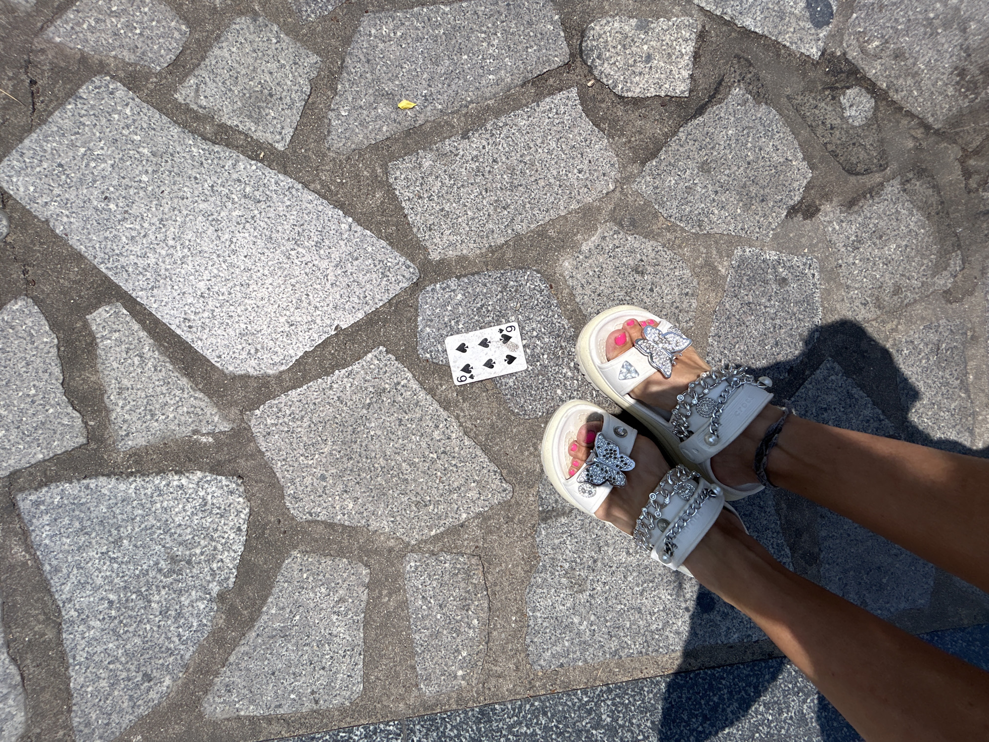 The image shows a section of paved stone ground, likely a sidewalk or plaza. A playing card, the 6 of spades, lies on the ground. A pair of female legs wearing white sandals adorned with silver butterfly embellishments and chains is visible in the bottom right corner. The location cannot be confirmed as Nha Trang, Vietnam, based on visual data alone.